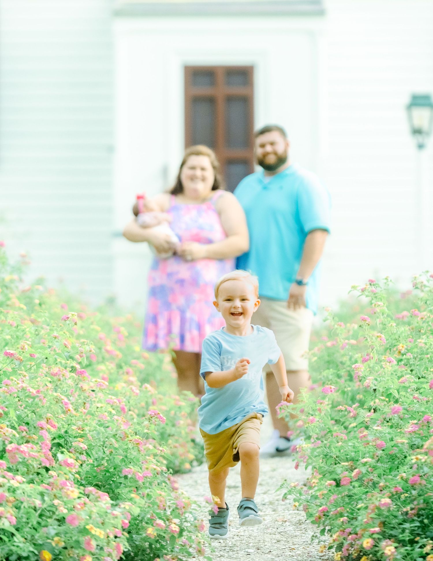 Children running and playing together through a garden path lined with pink flowering bushes on a bright sunny day.