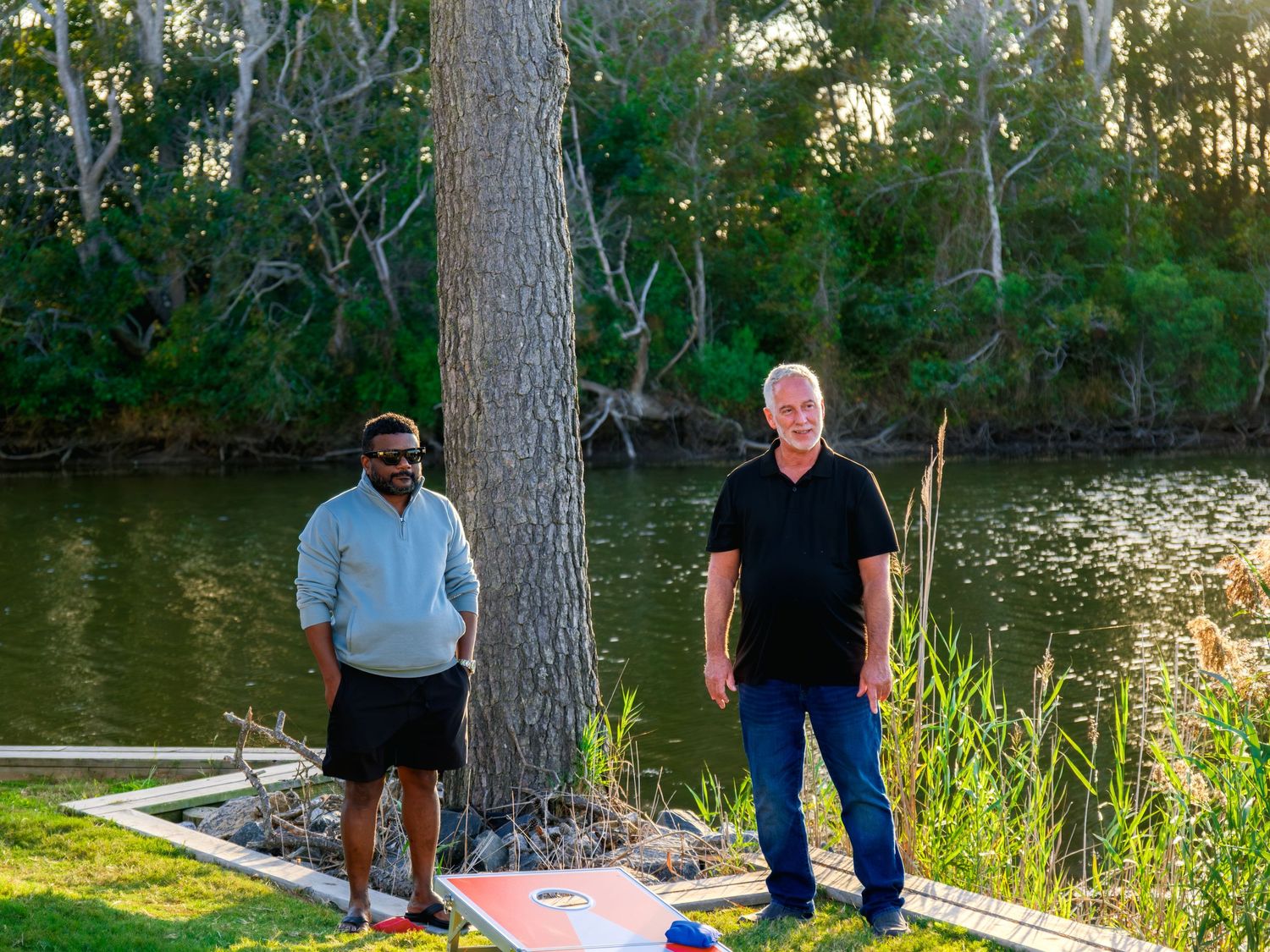 Two people standing beside a large tree trunk at the edge of a tranquil pond during golden hour.