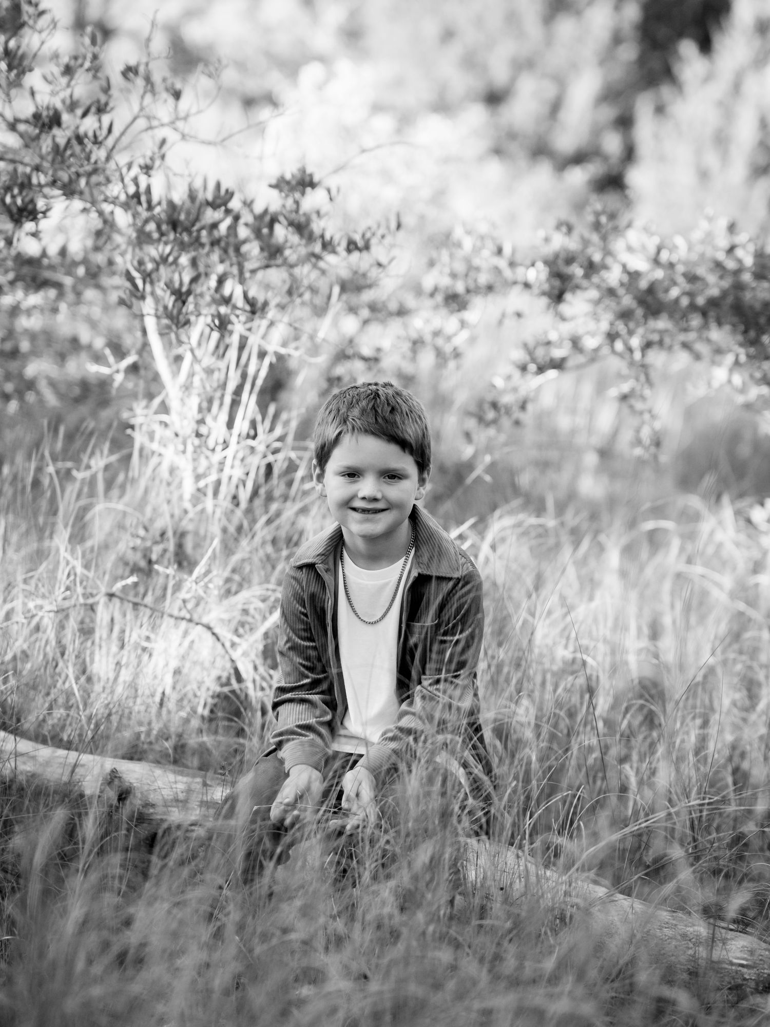 Black and white portrait photograph of a child sitting in tall grass with a gentle smile and wearing a casual jacket.