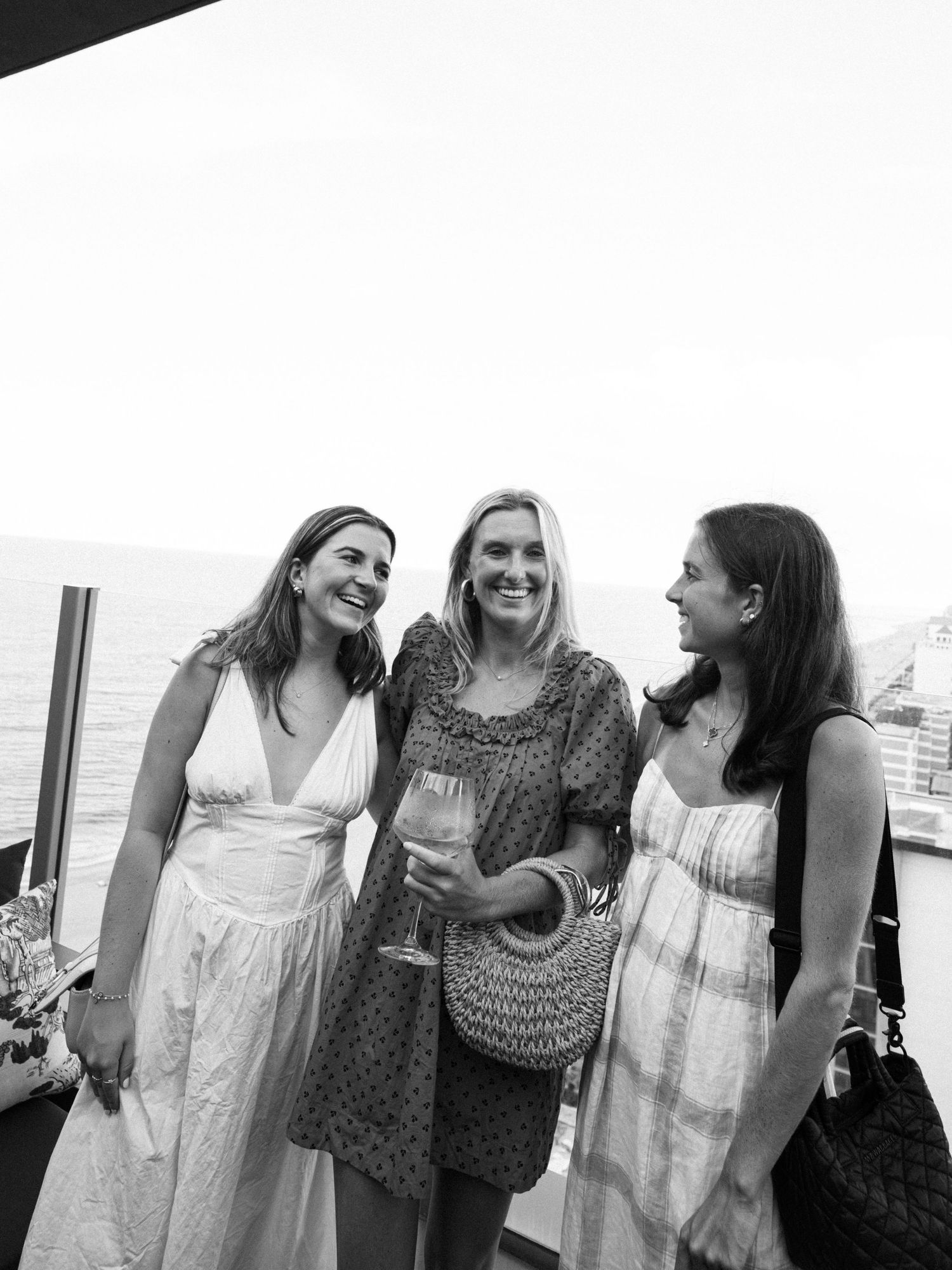 Three friends share a candid moment outdoors, laughing together while wearing summer dresses in black and white photography.