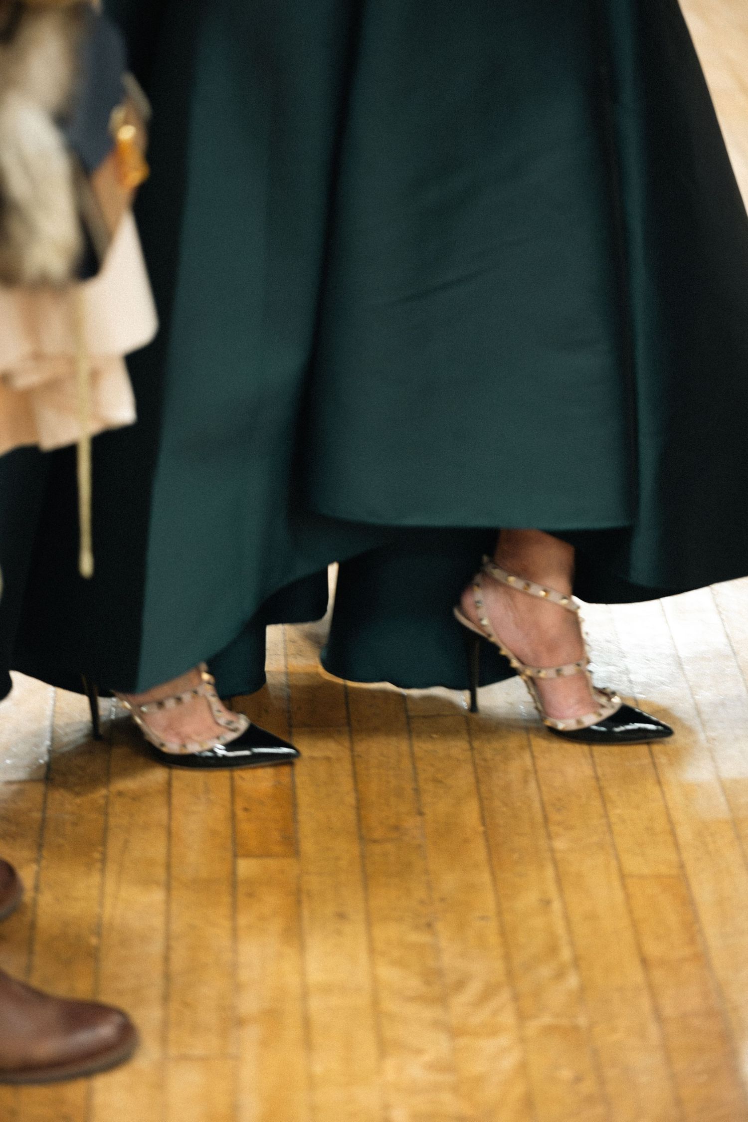 Close-up view of feet in black pointed flats with studded straps on wooden floor beneath a dark green skirt.