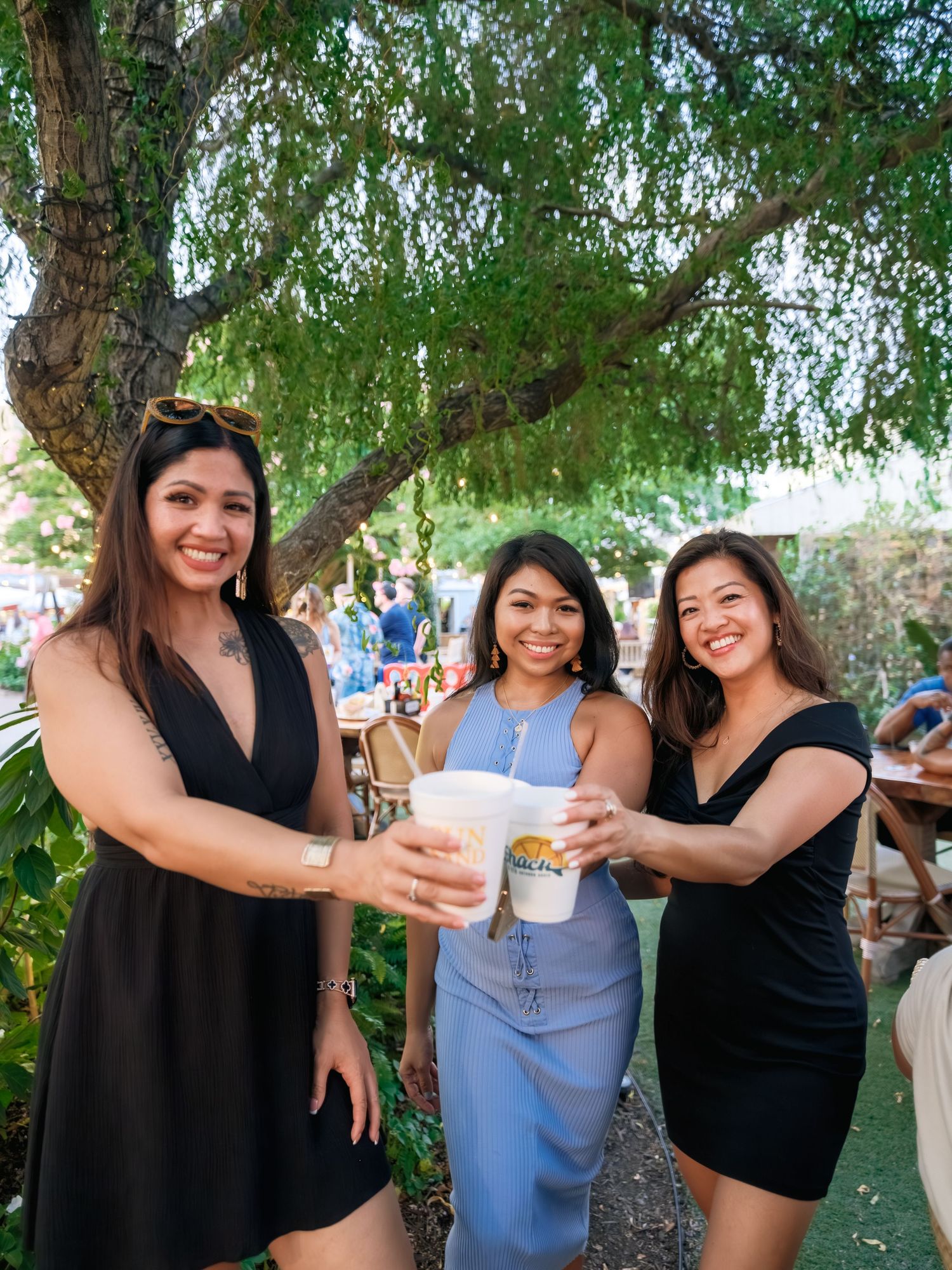 Three friends in dresses toast with drinks under a willow tree at an outdoor gathering.
