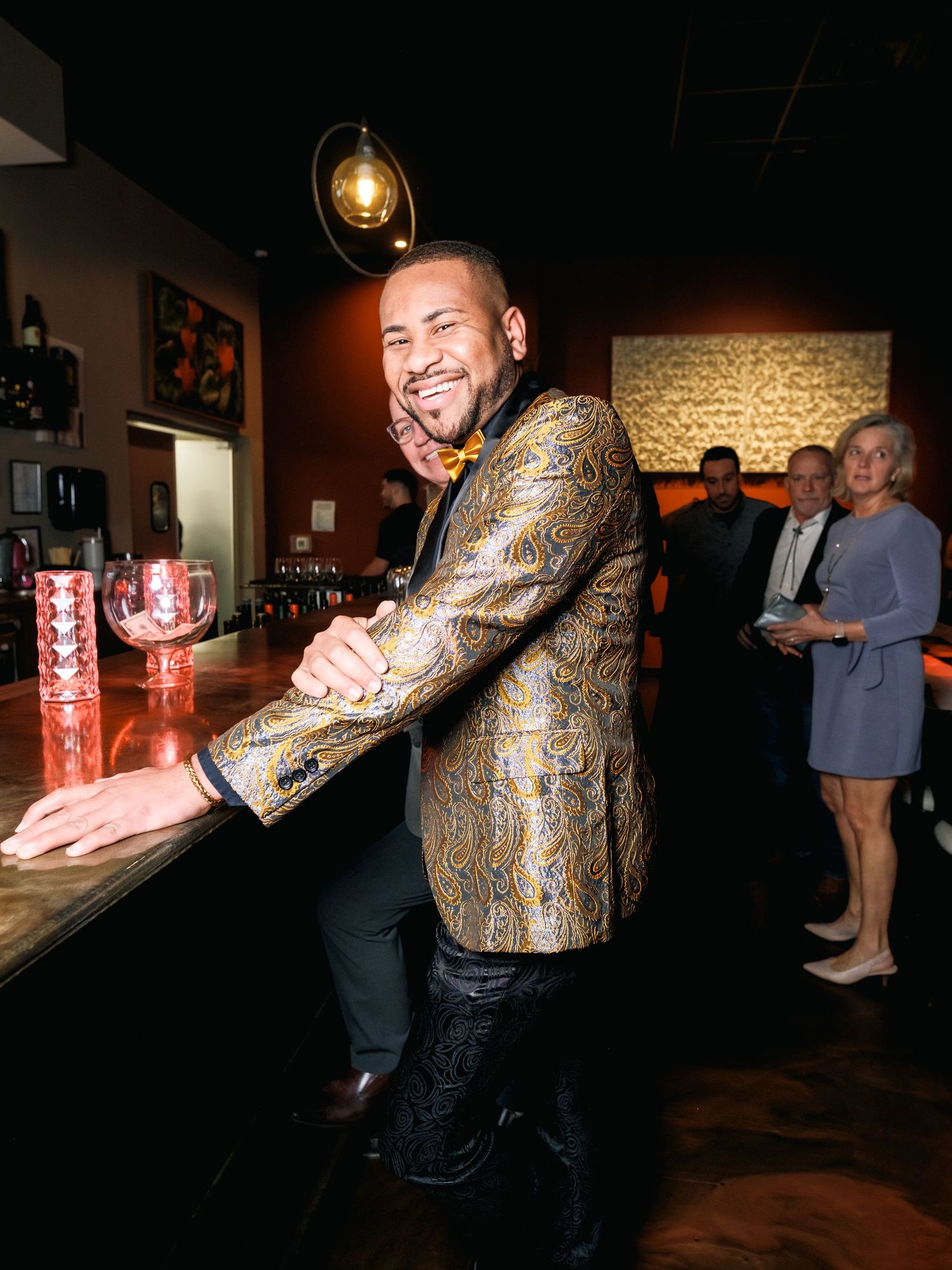 A person in a metallic patterned blazer smiles at a bar with guests in the background.