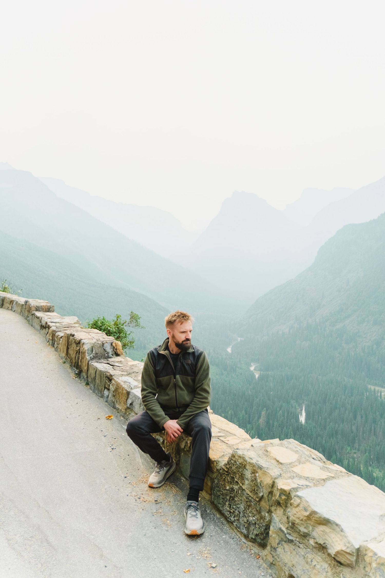 Person sits on stone wall overlooking misty mountain ranges and forest valley at Going-to-the-Sun Road in Glacier National Park.