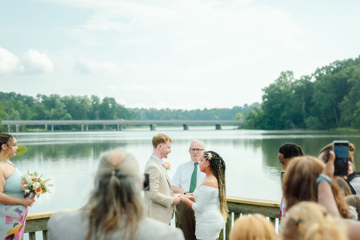 An outdoor wedding ceremony takes place on a wooden dock overlooking a serene lake surrounded by trees.
