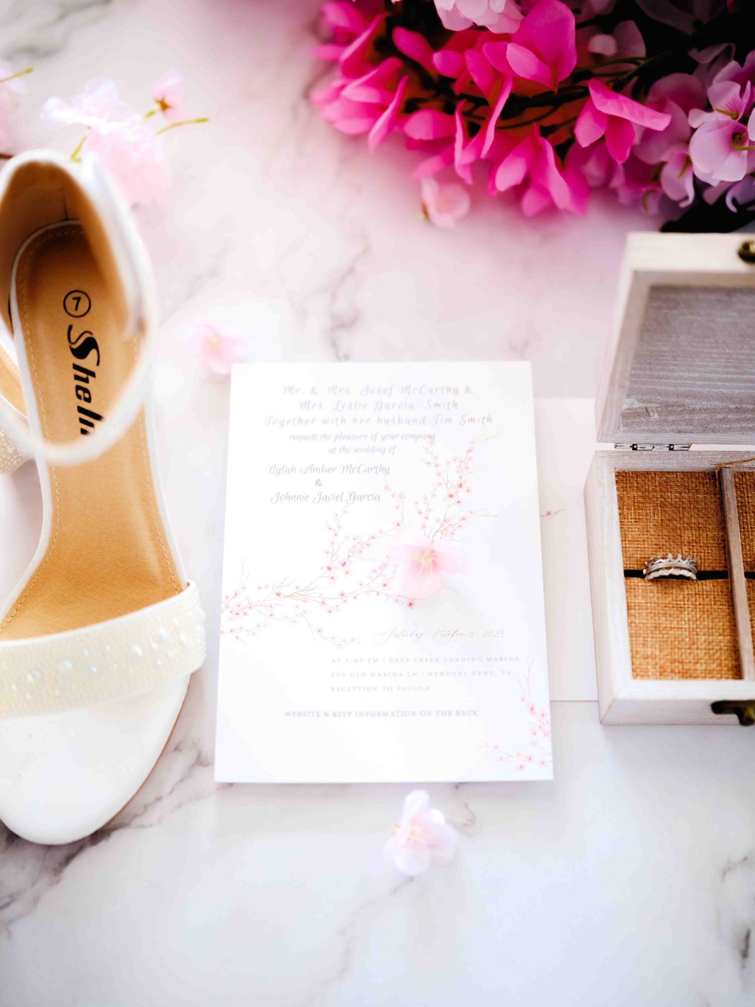 Wedding details flatlay with white bridal shoe, pink flowers, invitation and engagement ring box on marble surface.