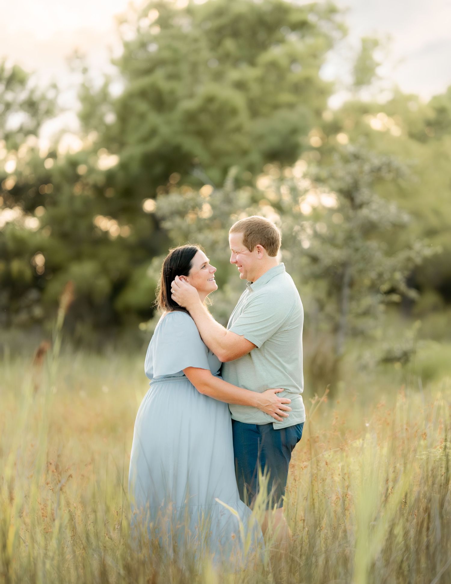A couple shares a tender embrace in a sunlit meadow wearing light blue outfits during golden hour.