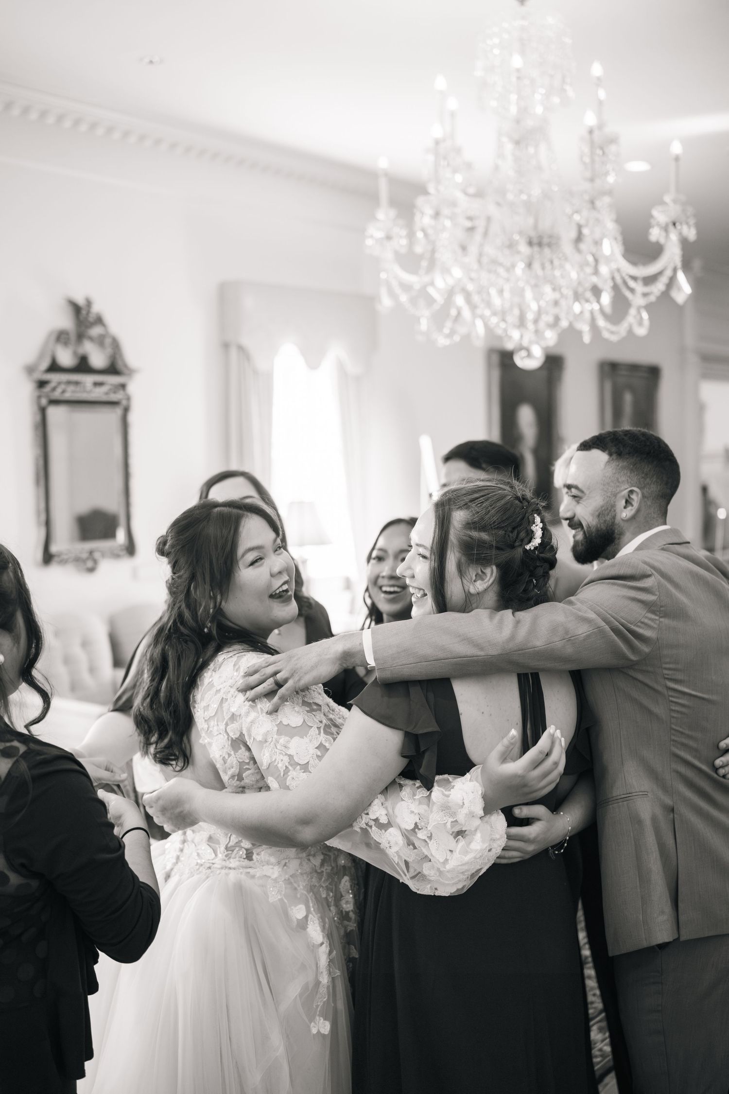 Guests embrace and laugh together during a celebration under elegant crystal chandeliers in a bright white room.