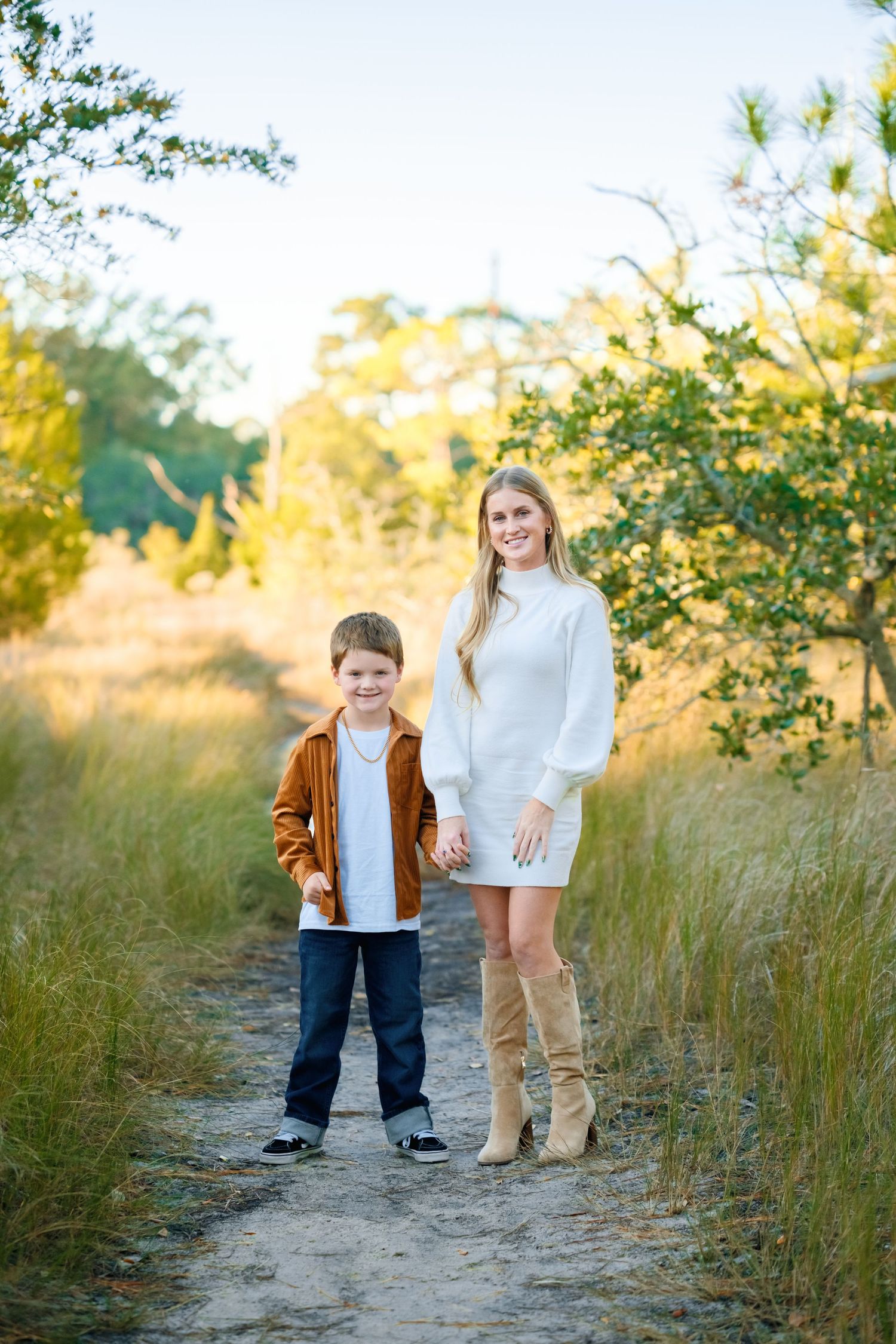 Family enjoying a golden sunset walk together on a sandy path through tall grass and trees.