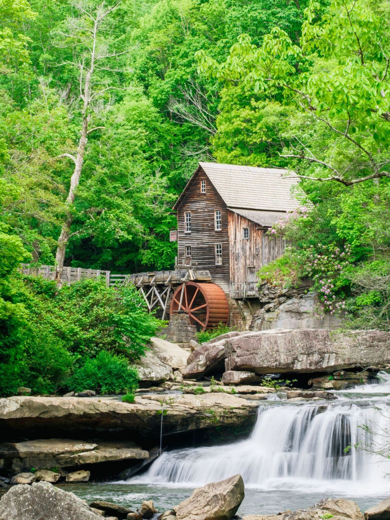 Historic wooden grist mill with water wheel sits beside cascading waterfall surrounded by lush green forest.