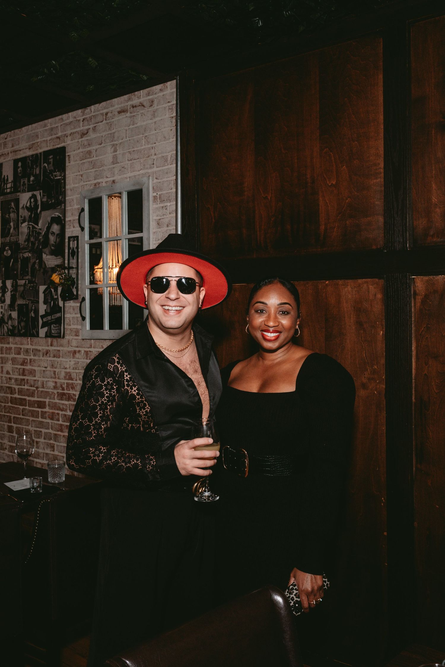 Two people stand together at a party venue with brick walls and dark wood paneling in the background.