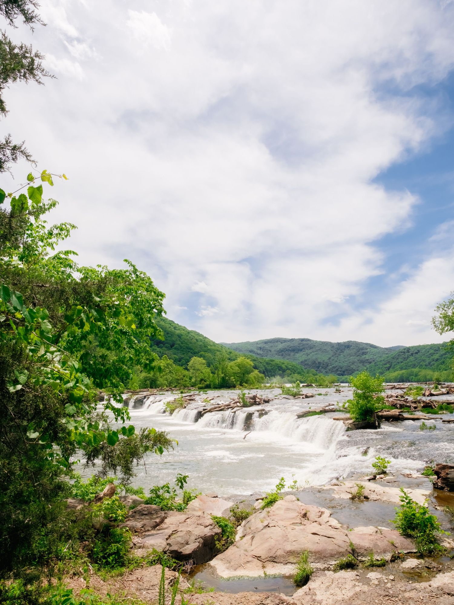 Scenic waterfall cascades over rocky terrain in a lush mountain landscape under cloudy summer skies.
