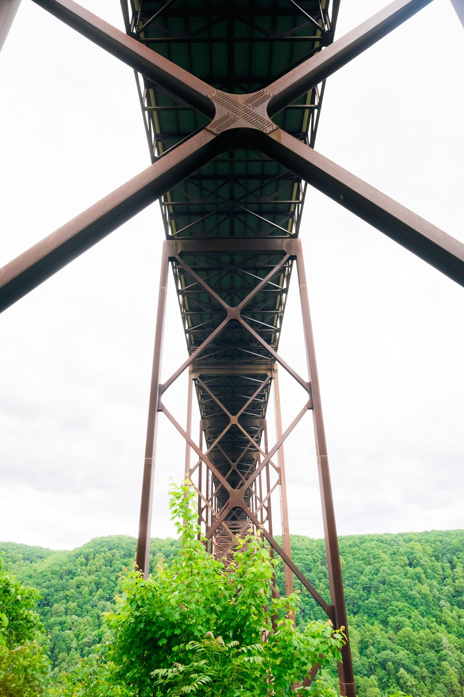 A dramatic upward view of the New River Gorge Bridge in West Virginia showing its massive steel support structure and green forest.