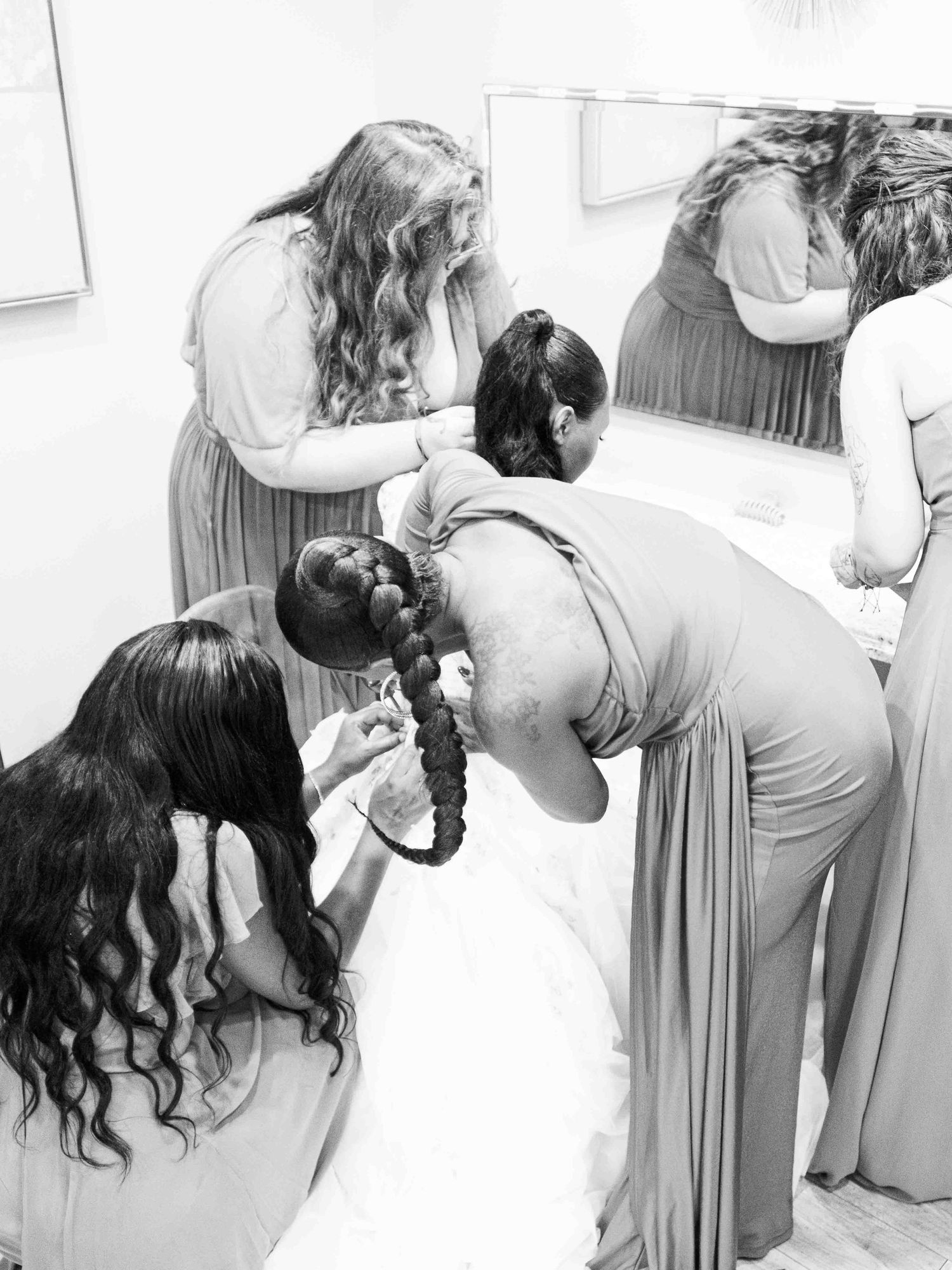 A black and white photo shows people in long dresses helping someone get ready, working together in a bridal prep moment.