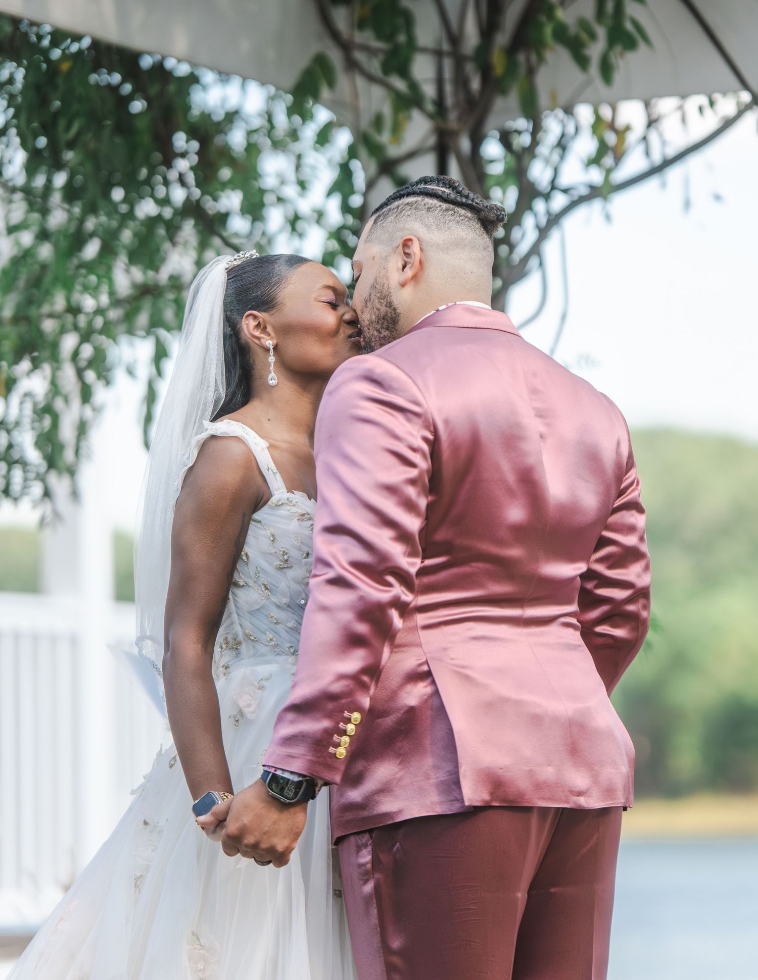 A tender moment between newlyweds in formal wedding attire, with the bride in white and groom in pink, sharing a kiss by a lake.