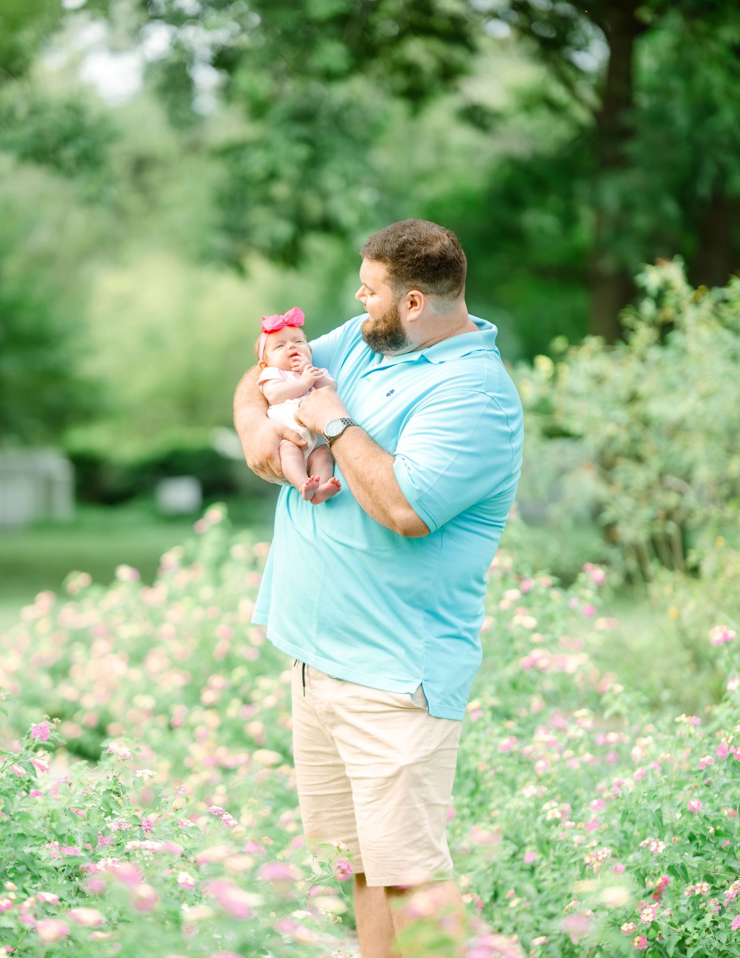 A caring moment outdoors with a small baby dressed in pink, surrounded by blooming flowers and lush greenery.