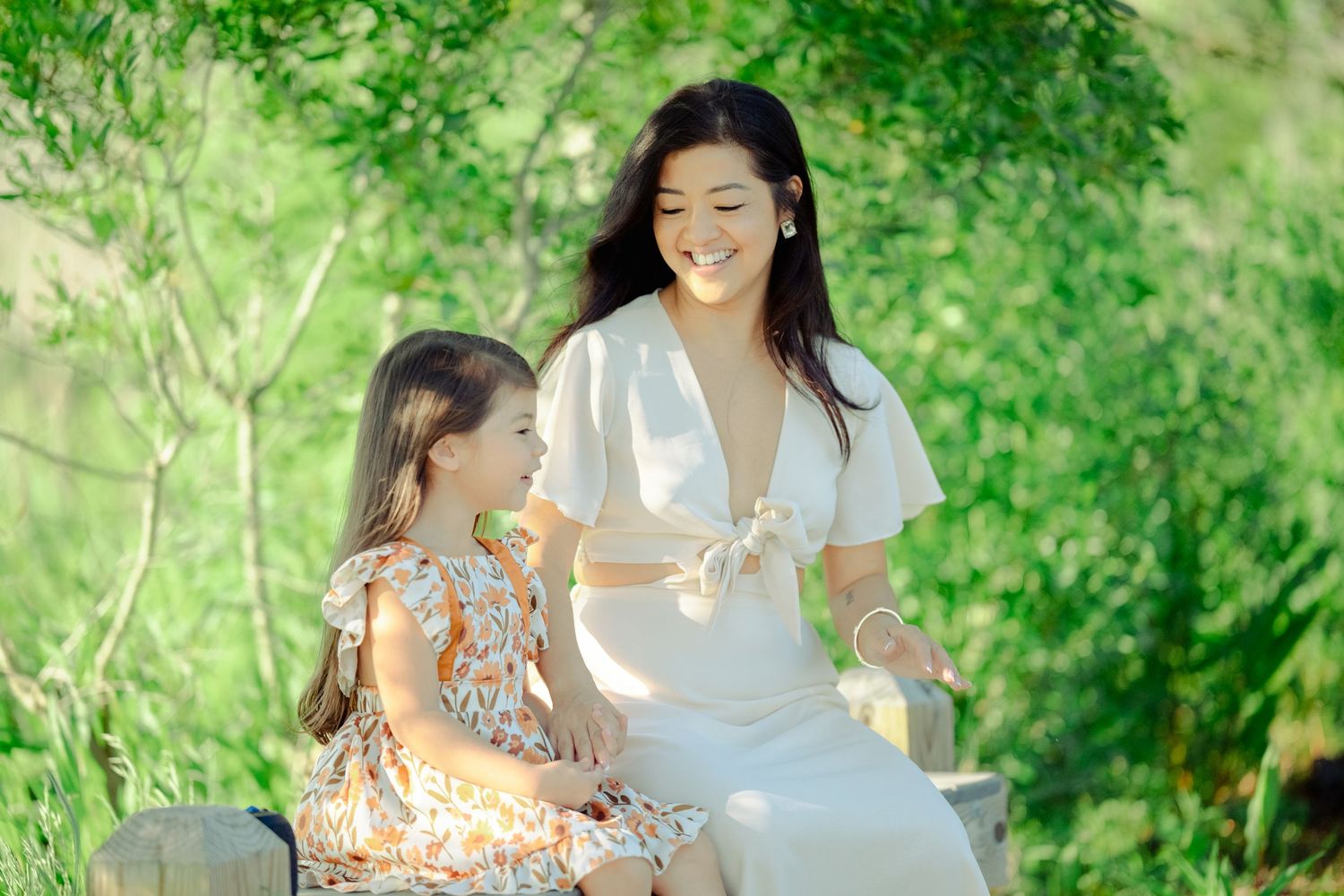 Two people in light summer outfits share a joyful moment sitting together outdoors against lush green foliage.