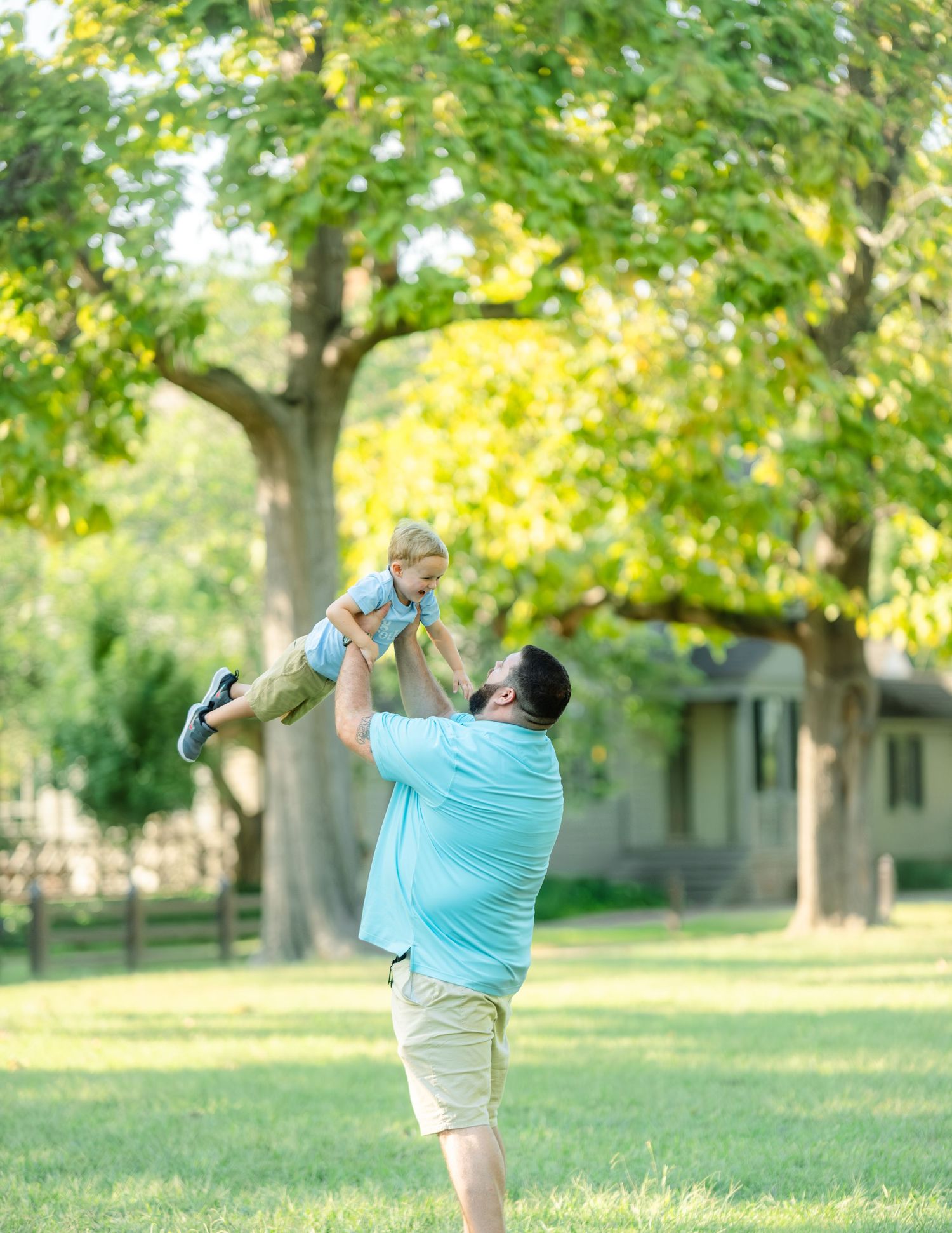 A person in a light blue shirt lifts a small child playfully into the air in a sunny park setting with green trees.