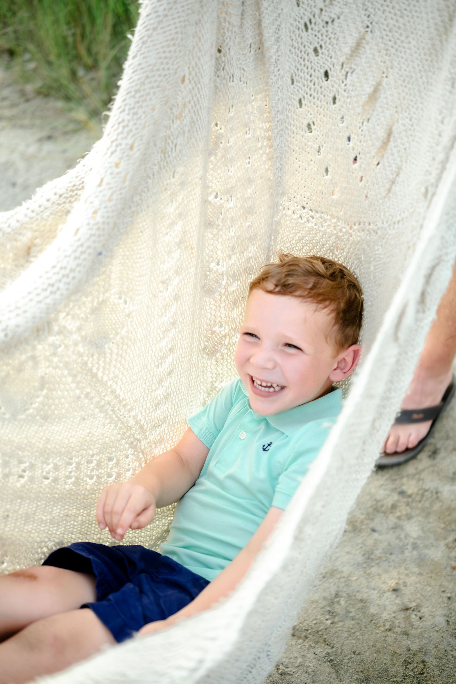 A figure in aqua colored clothing relaxes in a white hammock outdoors during summer.