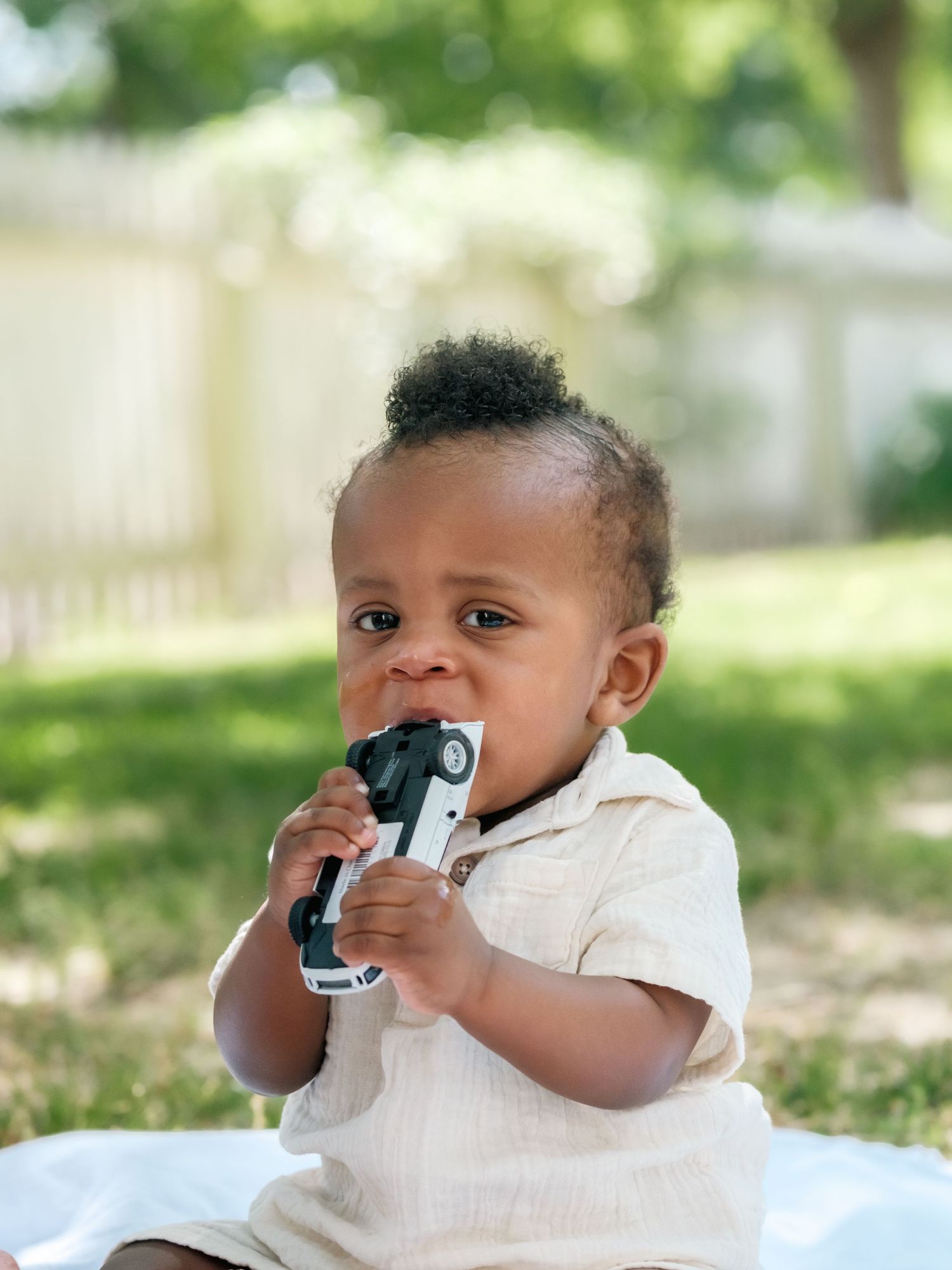 A baby in white clothing explores a digital camera with curiosity while sitting outdoors in a natural setting.