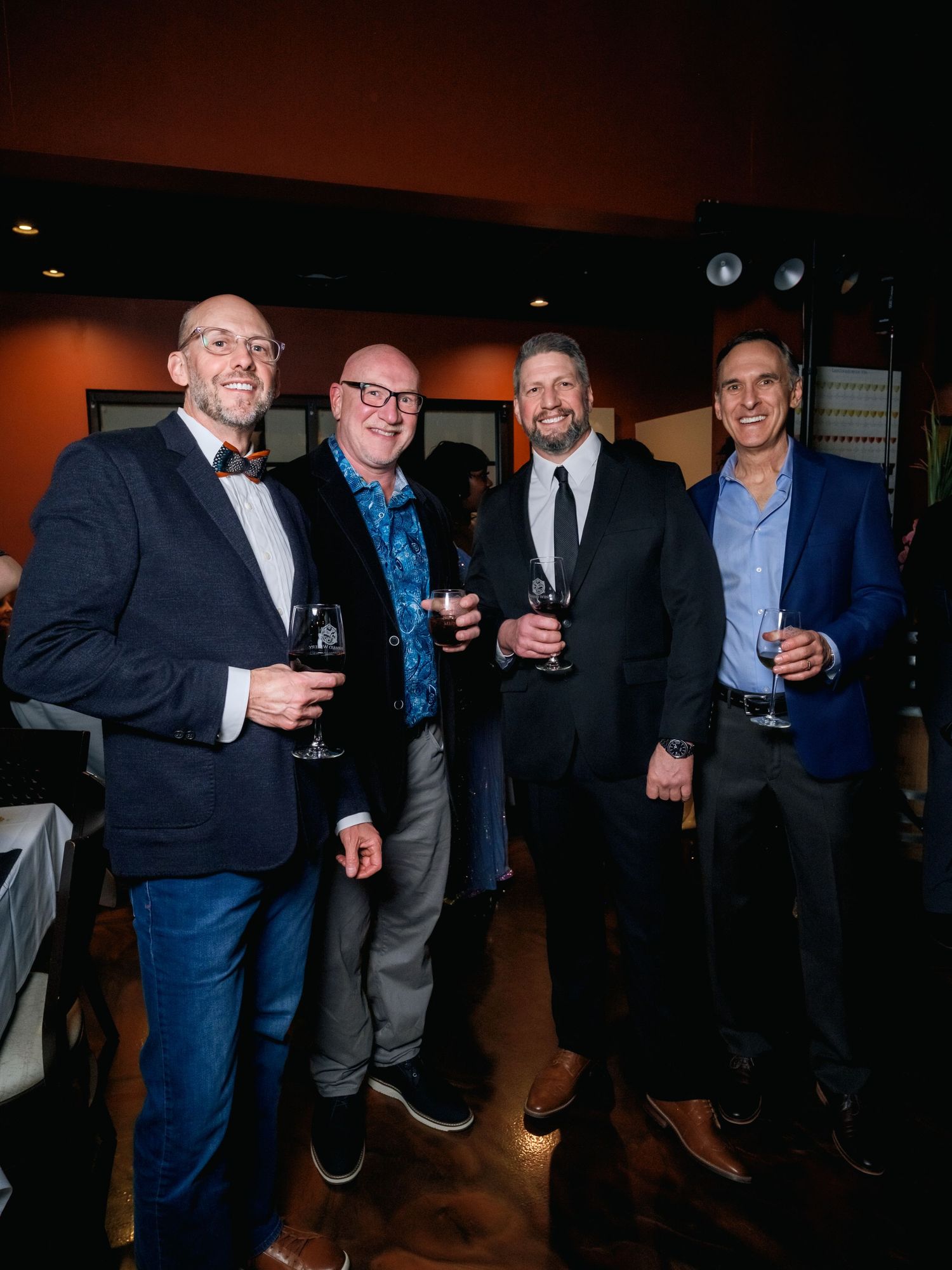Four men dressed in business casual attire smile while holding drinks at an evening social gathering.