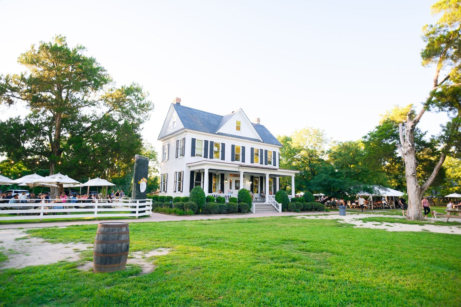 A panoramic view of a traditional white colonial-style house with manicured lawns and mature trees in daylight.