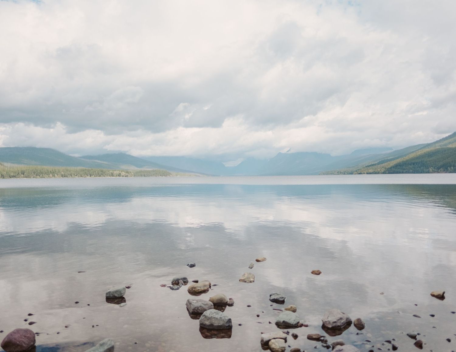 A panoramic view of a tranquil lake surrounded by mountains with scattered rocks along the shoreline in foggy conditions.