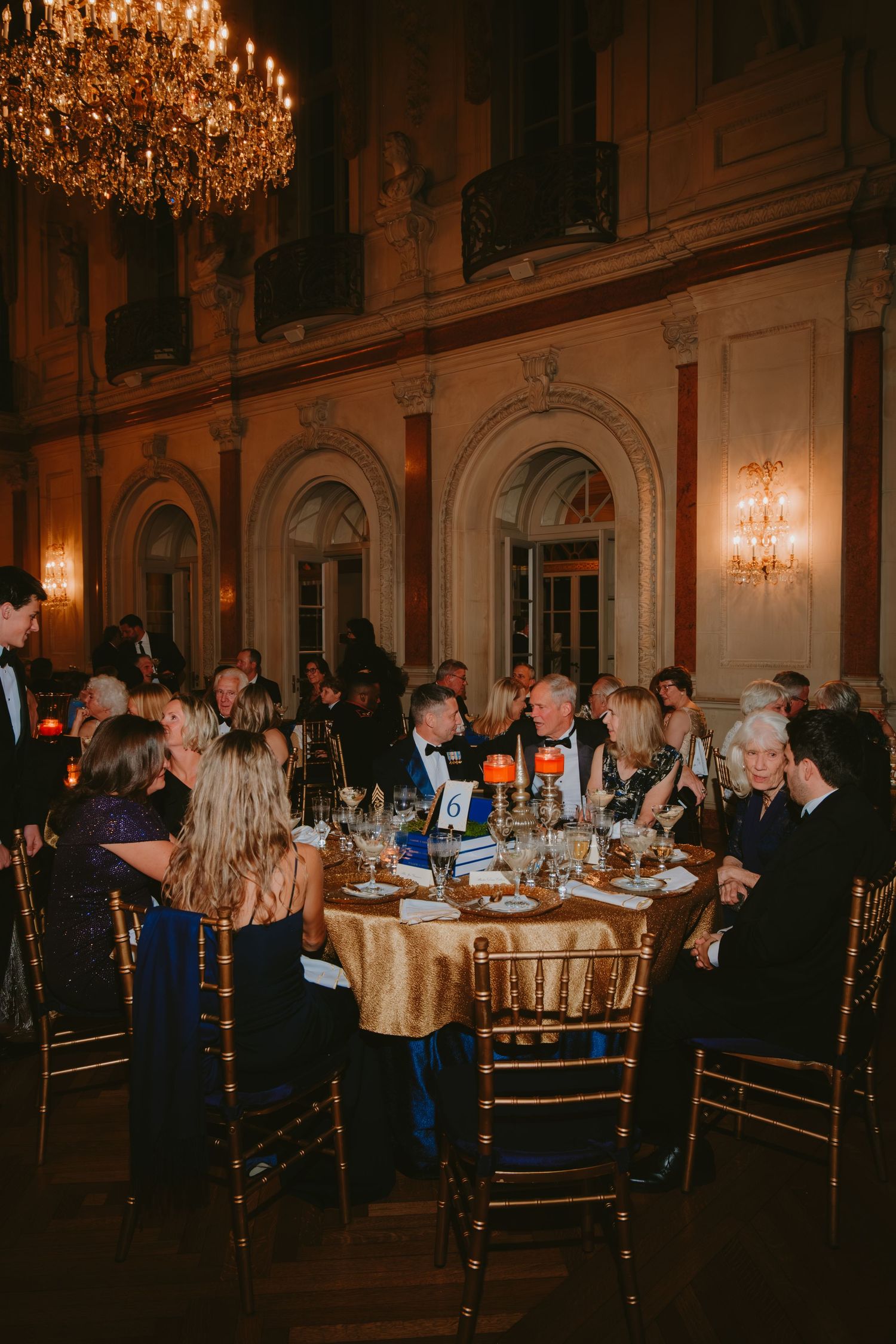 Guests seated at elegant round tables in an ornate ballroom with crystal chandeliers and gilded decor during an evening event.