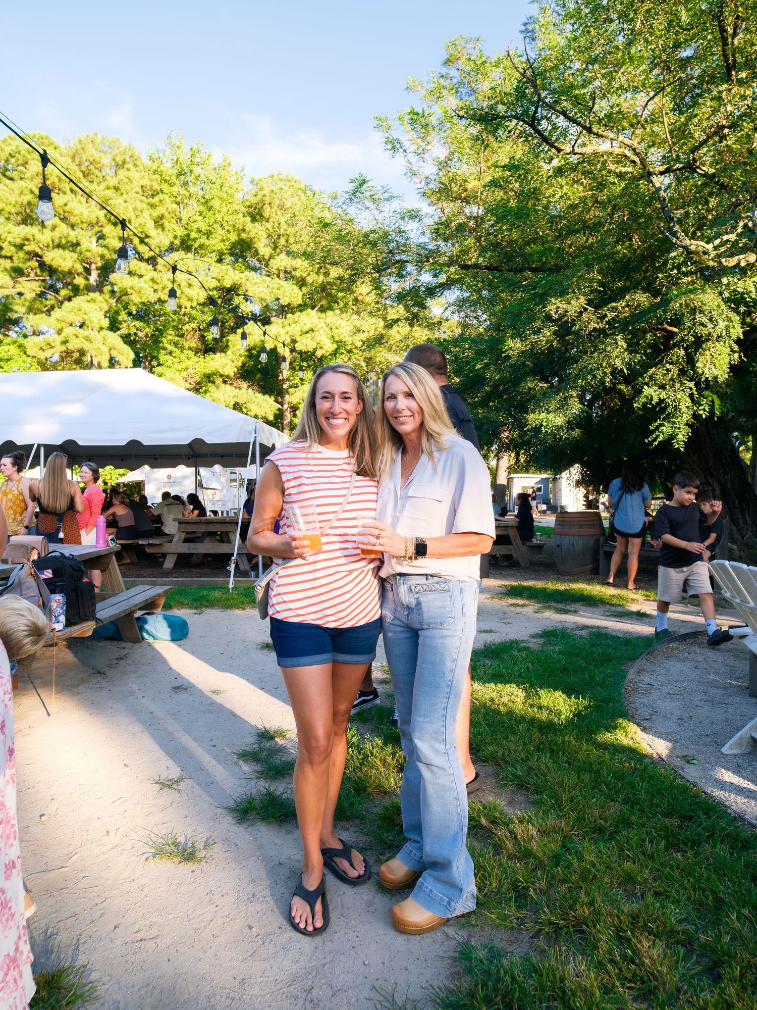 Visitors enjoy a summer outdoor gathering with tents and seating on a grassy lawn surrounded by leafy green trees.