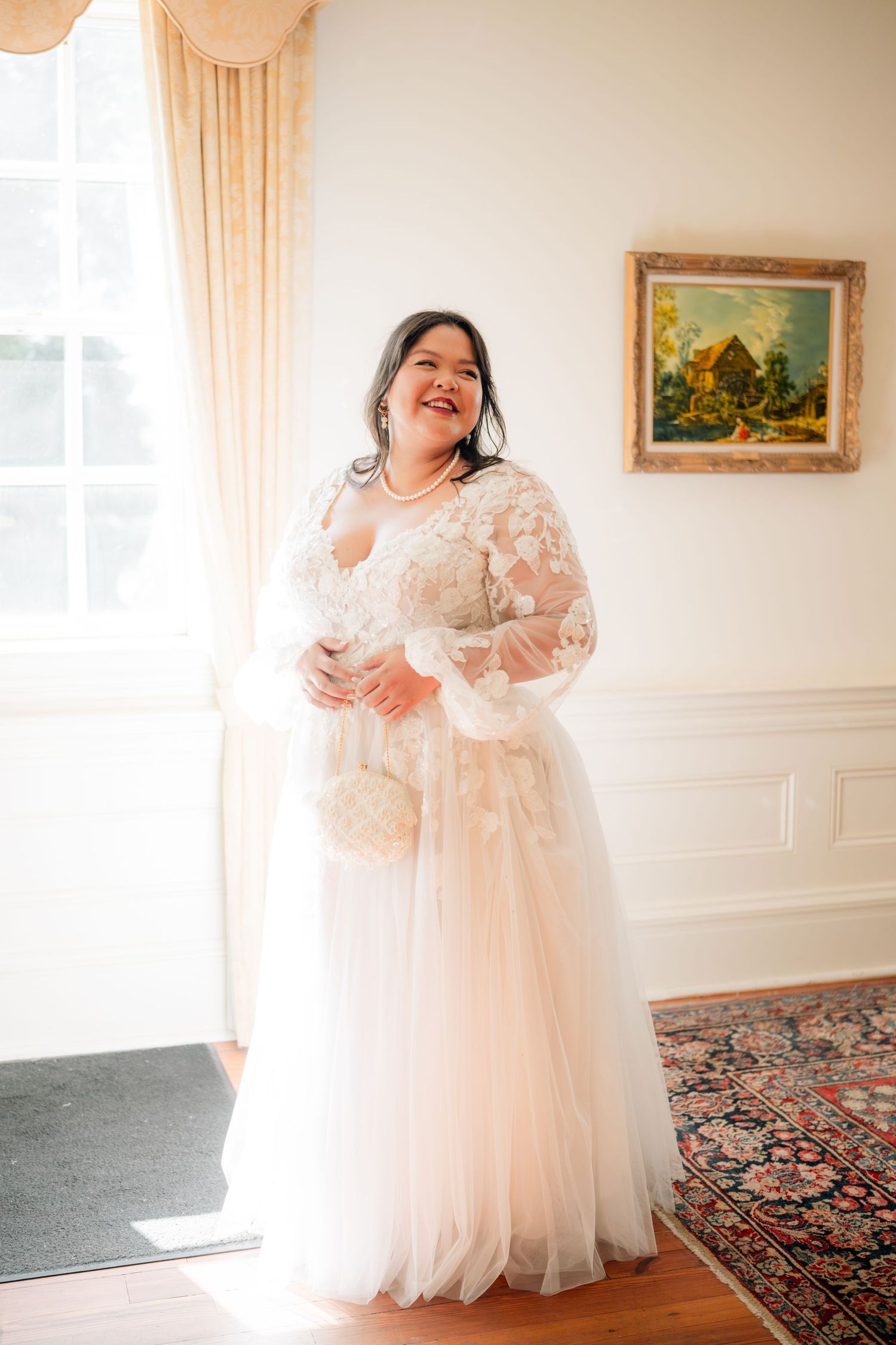 A bride wearing a romantic long-sleeved lace and tulle wedding gown stands in natural light by a window with peach curtains.