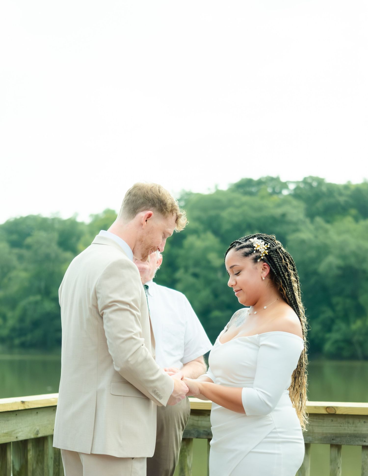 A wedding couple stands on a wooden deck by a lake exchanging rings during an intimate outdoor ceremony.