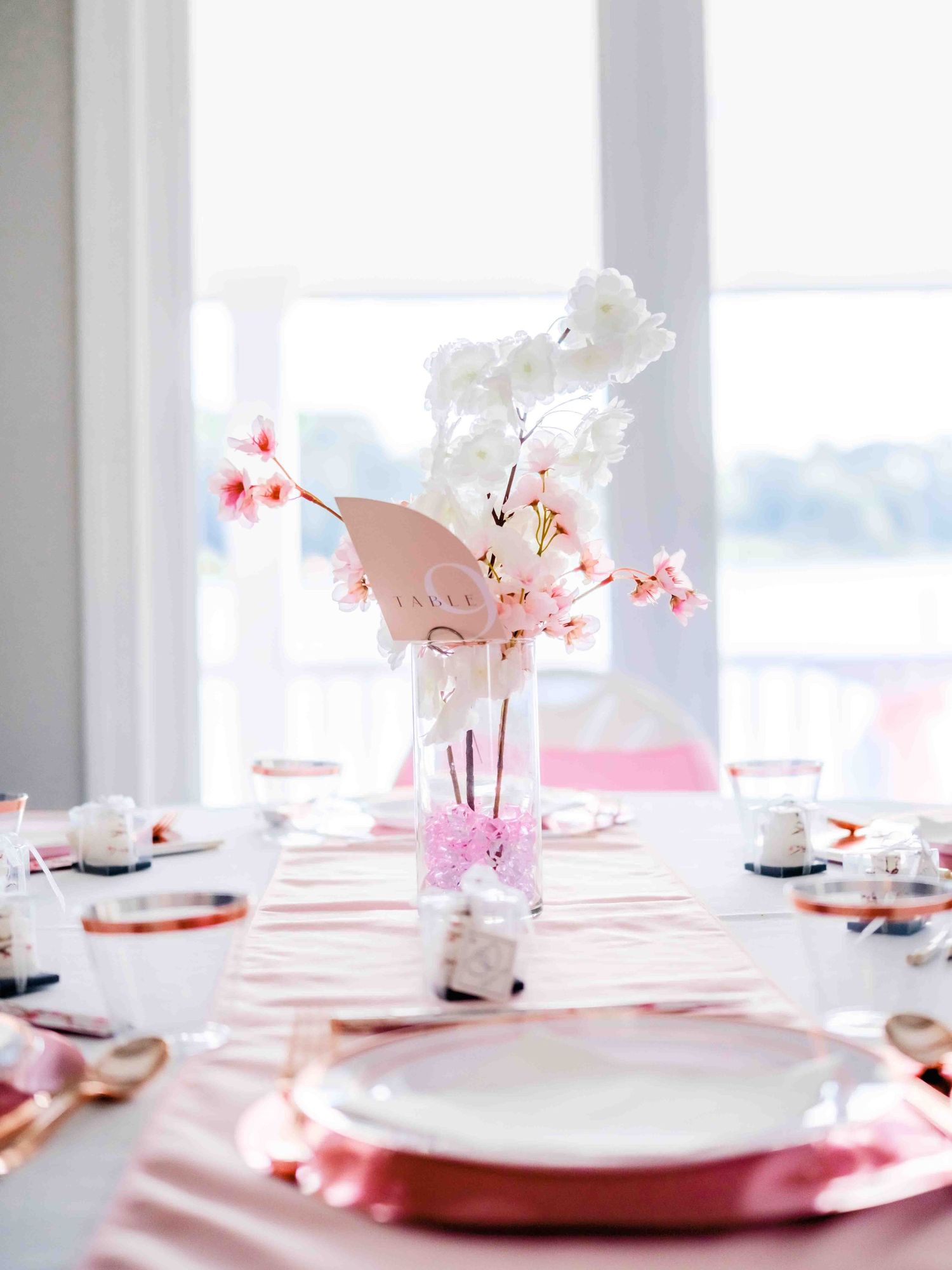An elegant table setting with pink charger plates, white dishes and delicate cherry blossom centerpieces in a bright room.