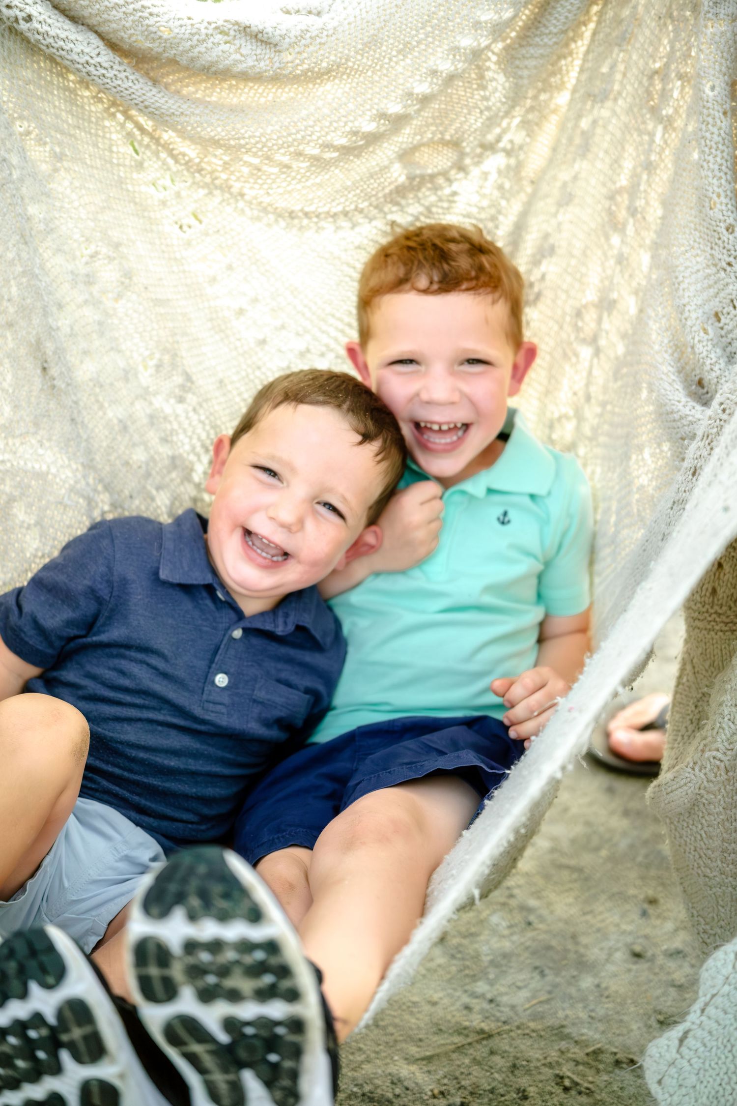 Two children in blue and teal shirts sit together on a hammock while laughing and smiling.