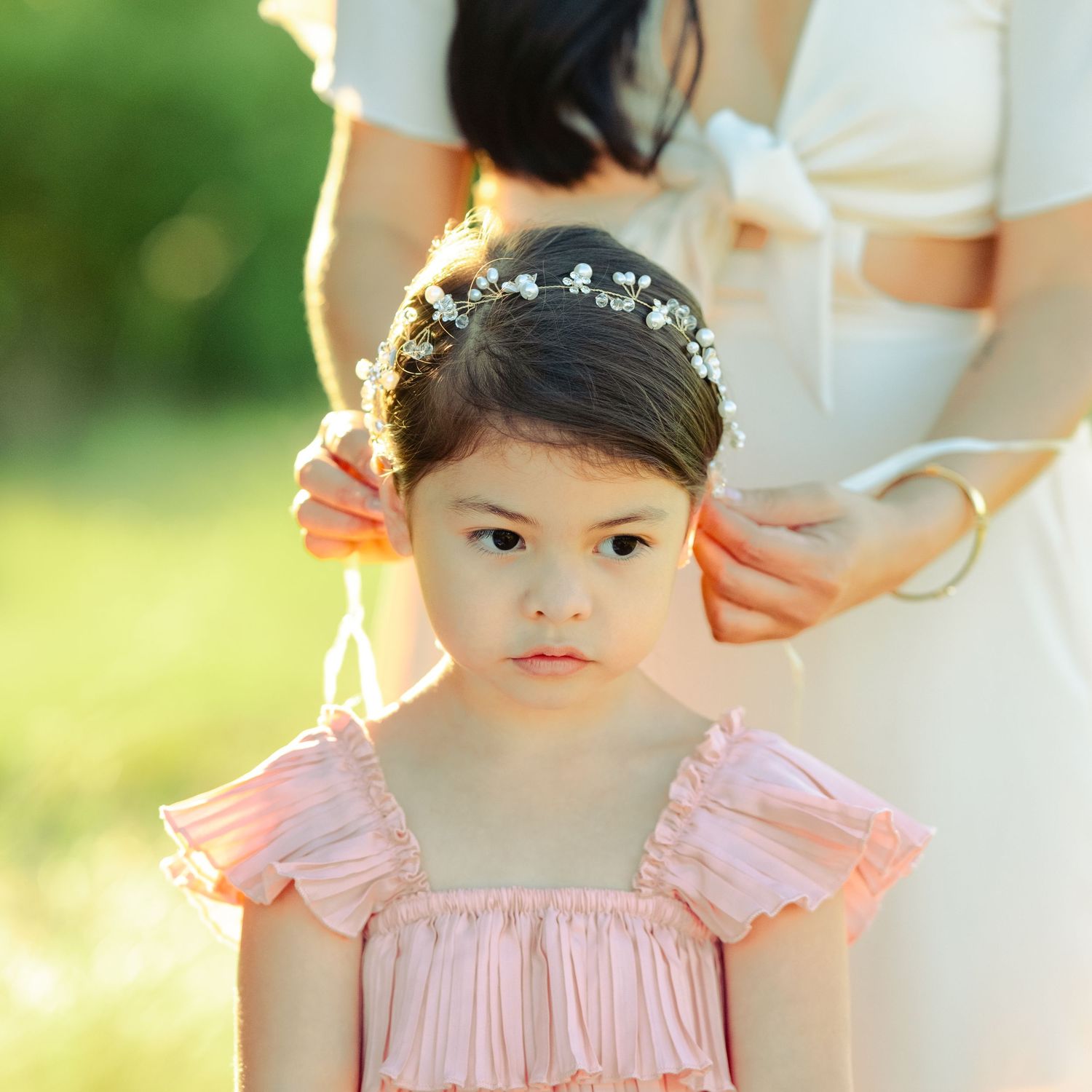 A child wearing a pink dress and floral headband stands in soft sunlit garden setting.