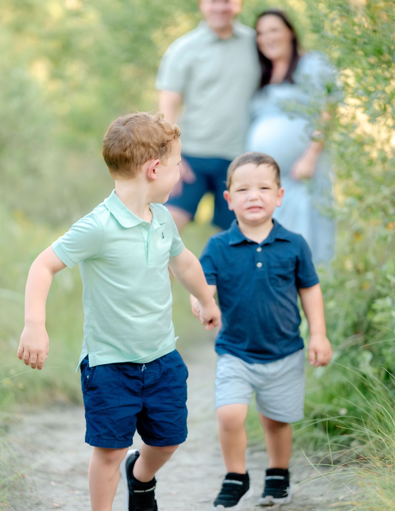 Two children in casual clothing running and playing on a nature trail with adults in the background.