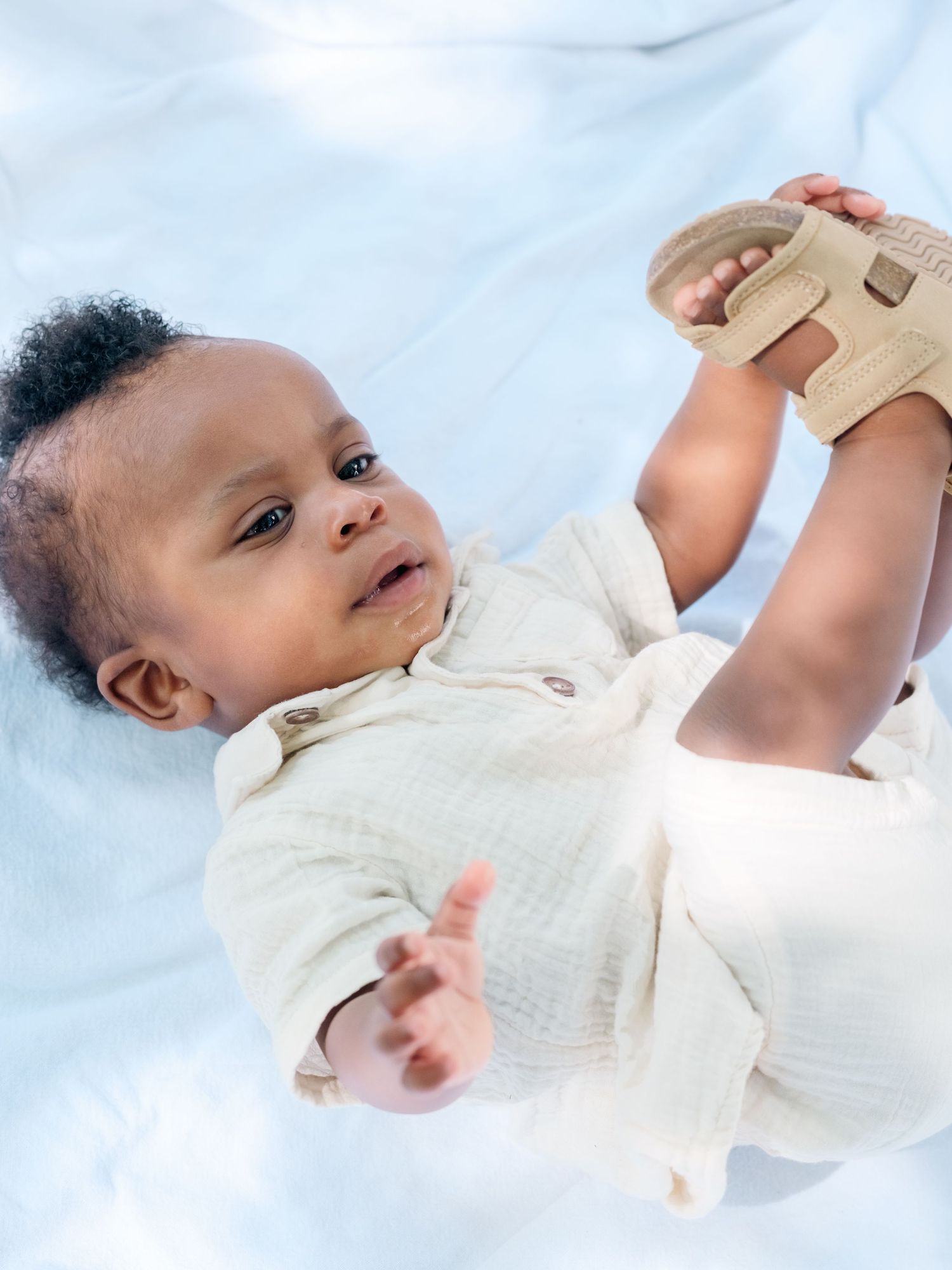 Small baby wearing a white outfit lies on soft blue fabric while holding and playing with a tan-colored toy.
