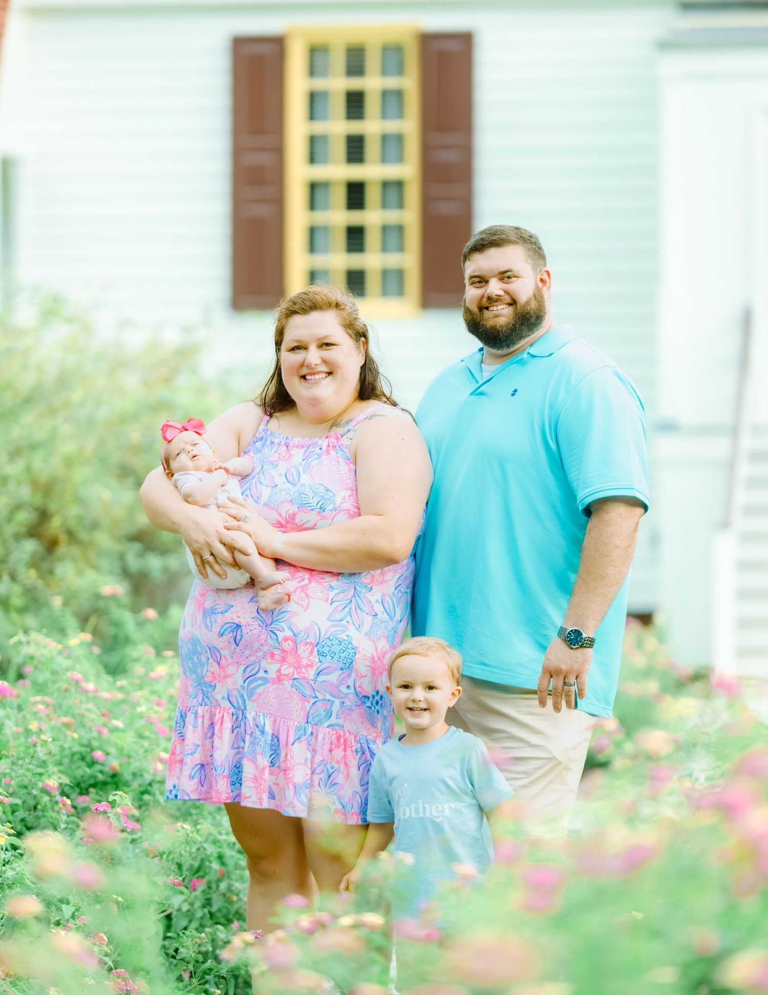 A couple in coordinating teal and pink outfits poses with a toddler in a blooming garden setting.