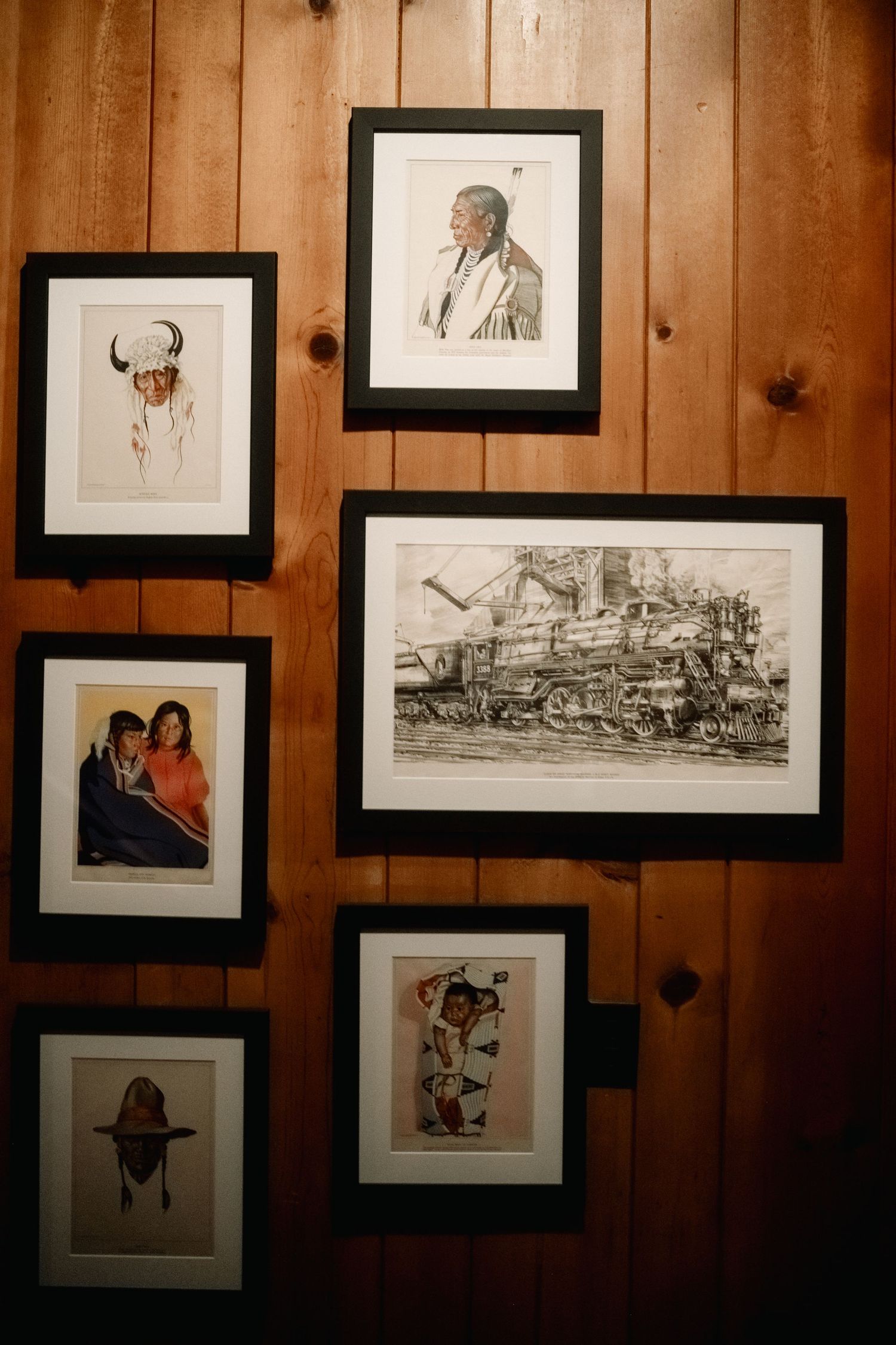 A collection of framed artwork and photographs arranged on a wooden paneled wall in gallery style.