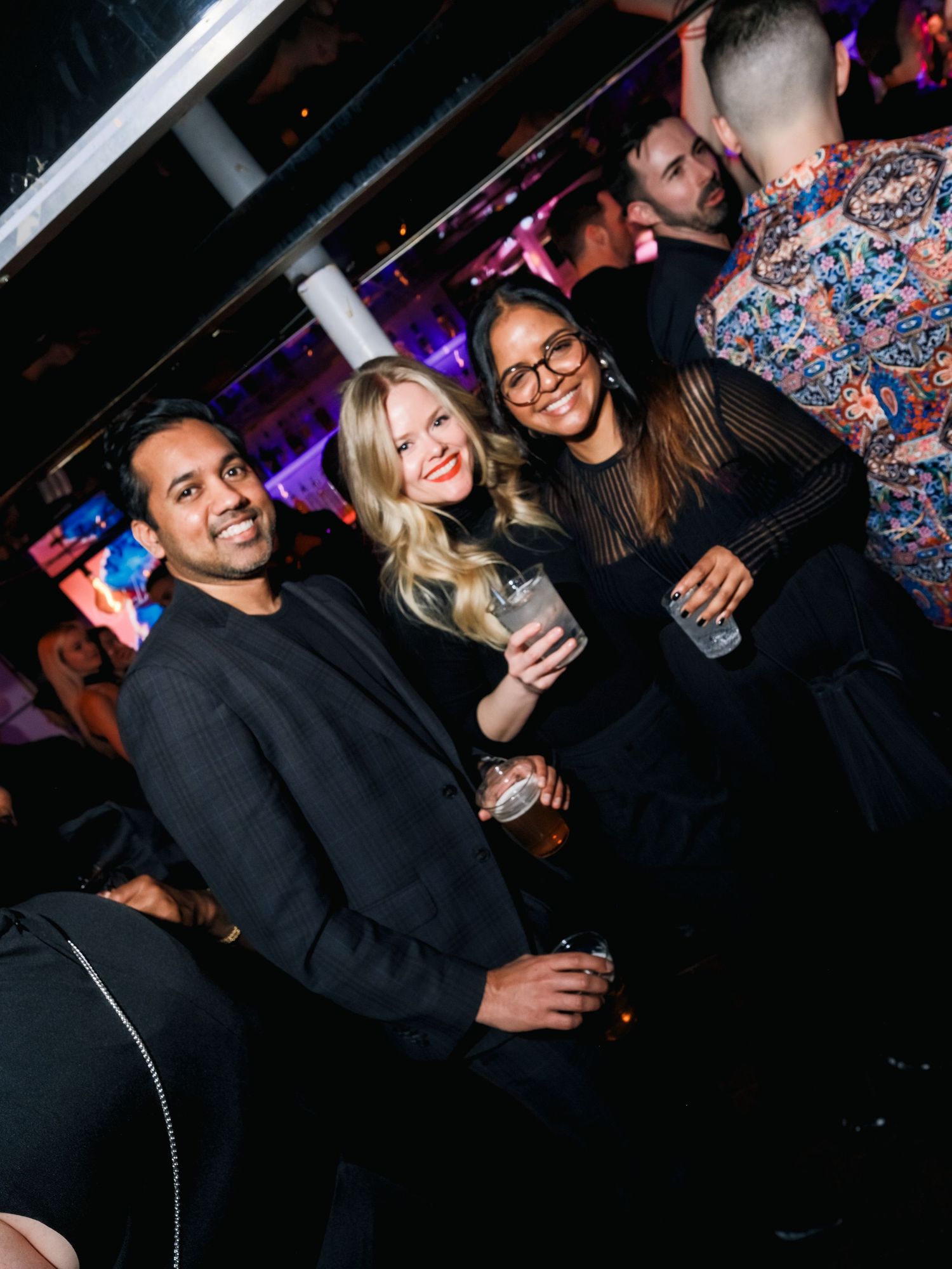 A group of friends enjoying drinks and socializing at a nightclub with purple lighting in the background.