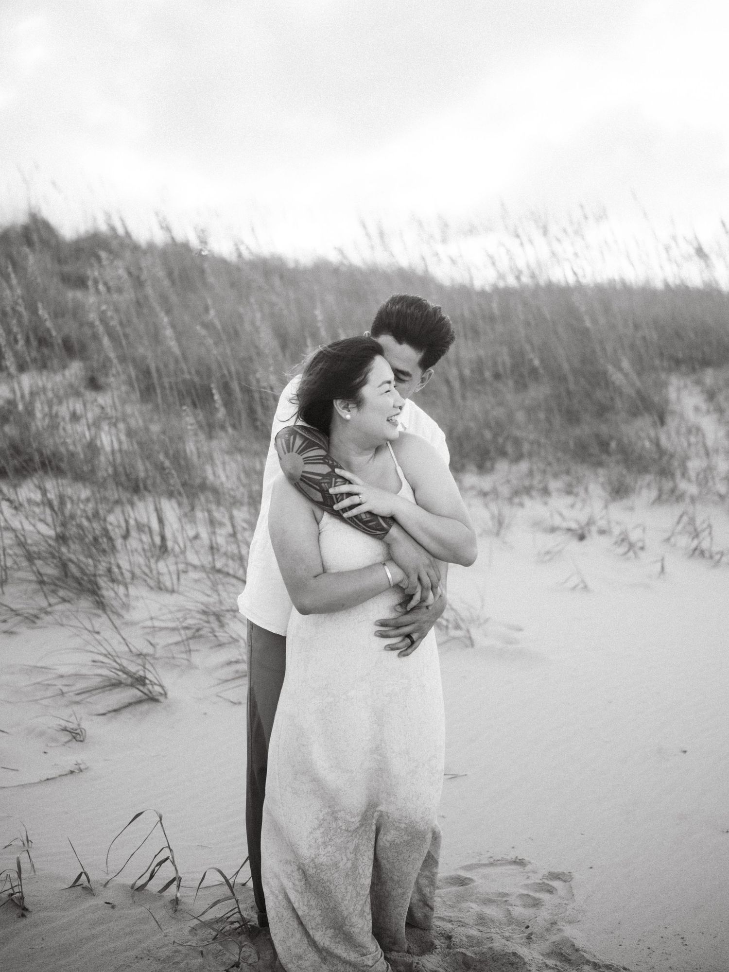 Black and white photo of couple sharing intimate moment among beach grass dunes.