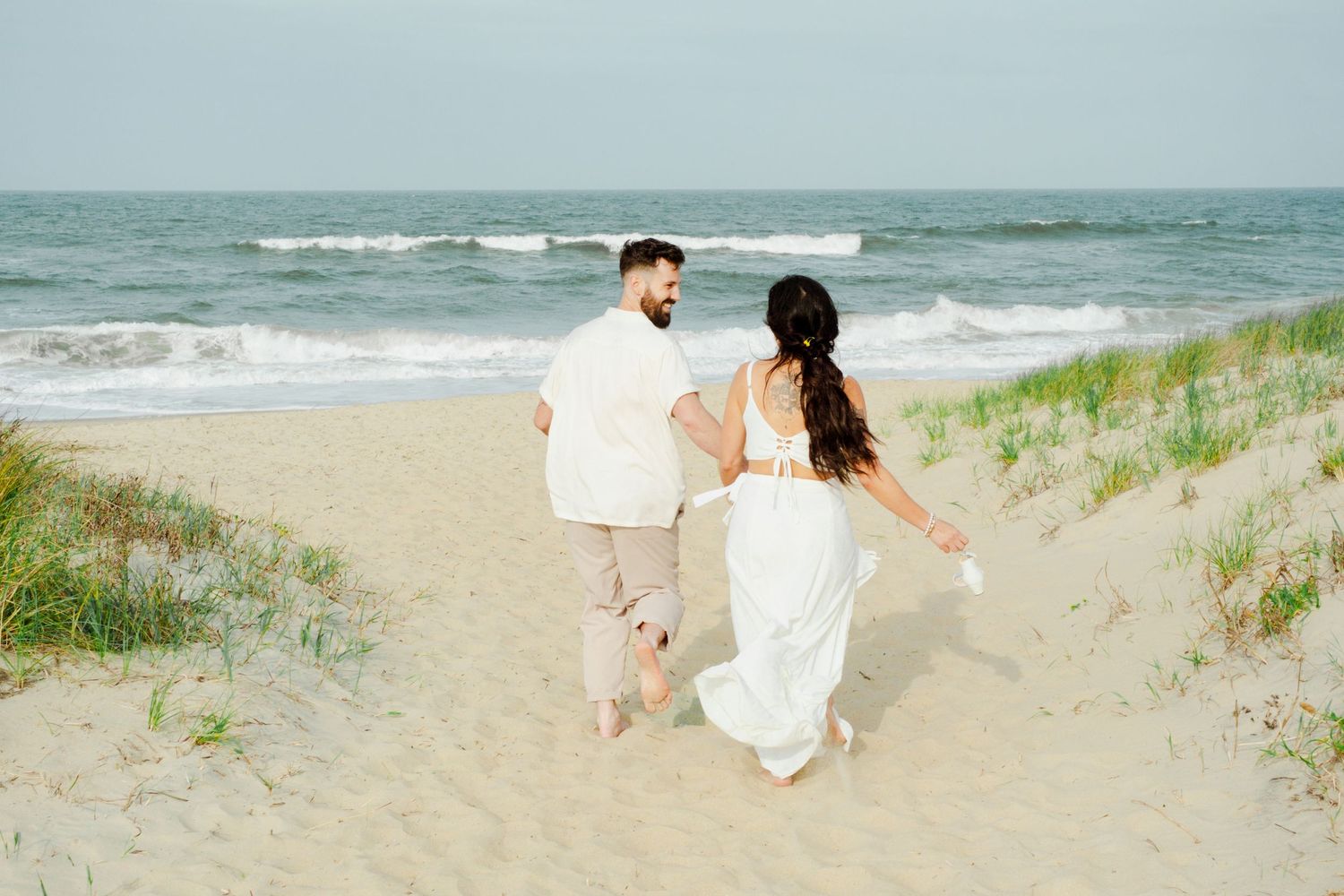 A couple walks hand in hand down a sandy beach path with ocean waves and dune grass in the background.