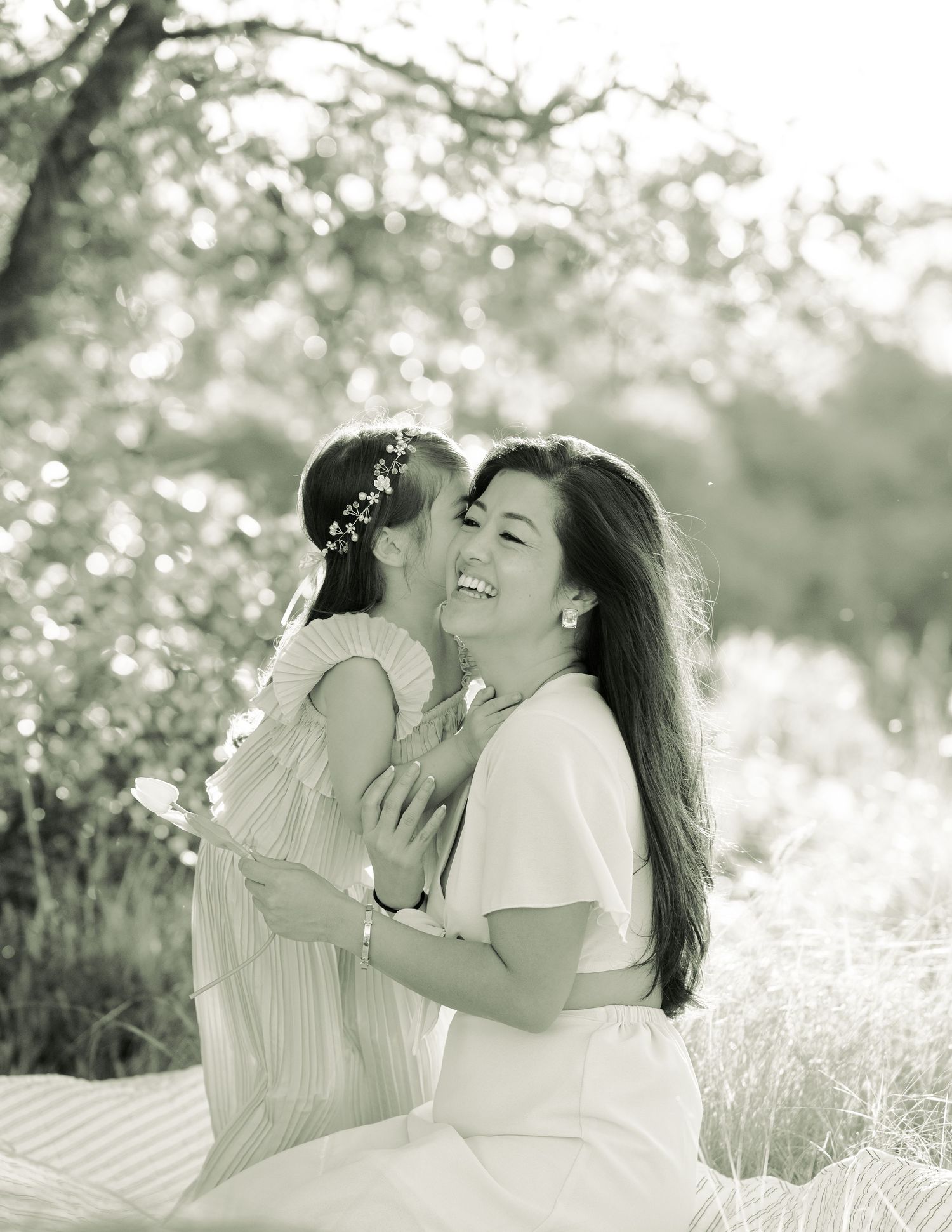 A tender black and white portrait of a parent and child sharing a joyful embrace in a sunlit outdoor setting.