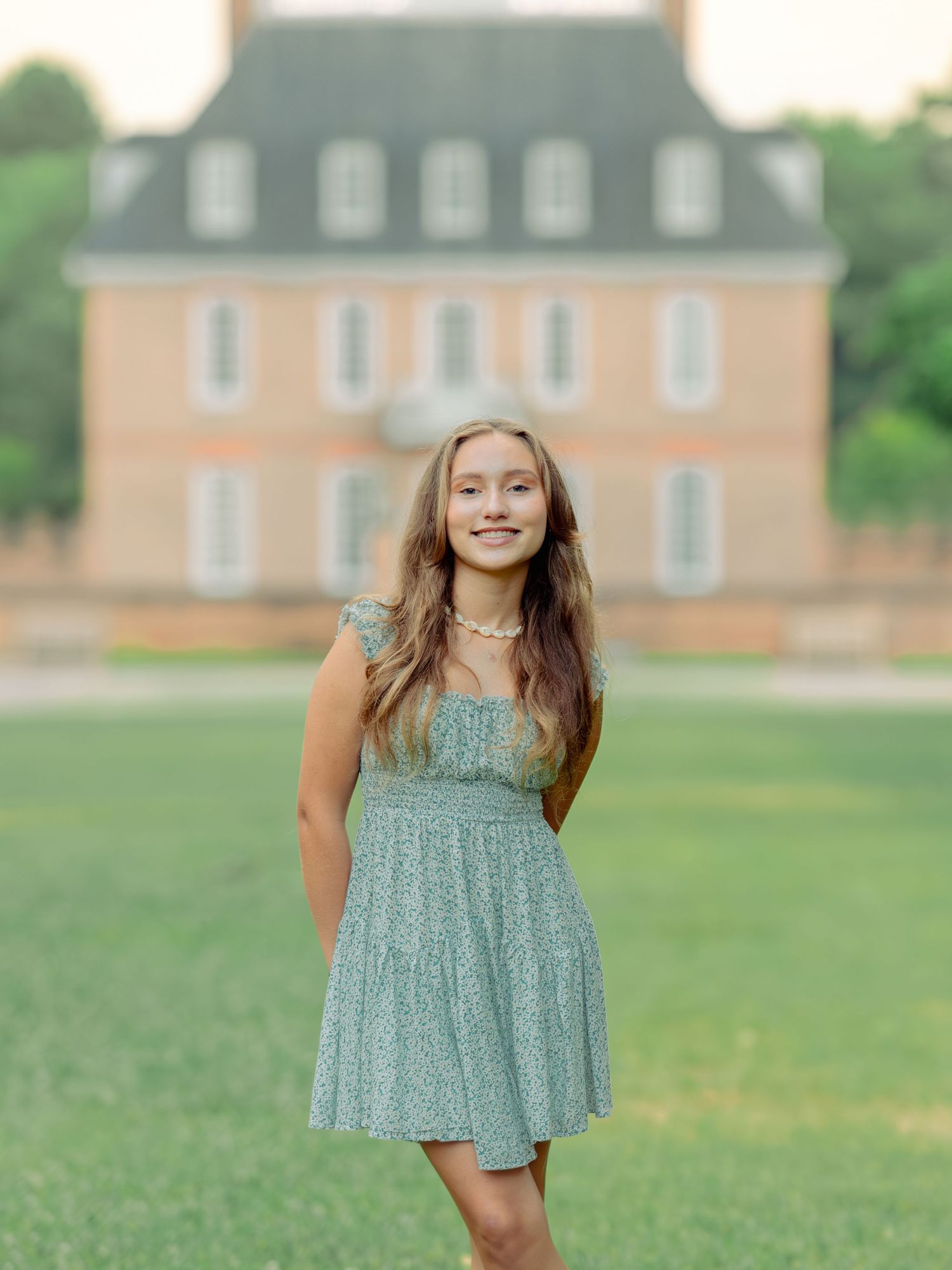 Someone wearing a casual sage green sundress poses on a lawn in front of a stately pink mansion with white trim.