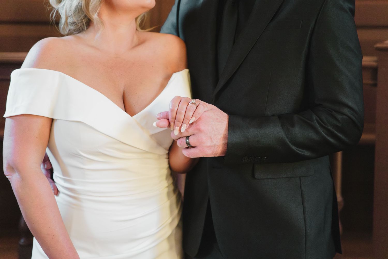 Close-up detail shot of a couple at their wedding, showing off-shoulder white gown and black suit.