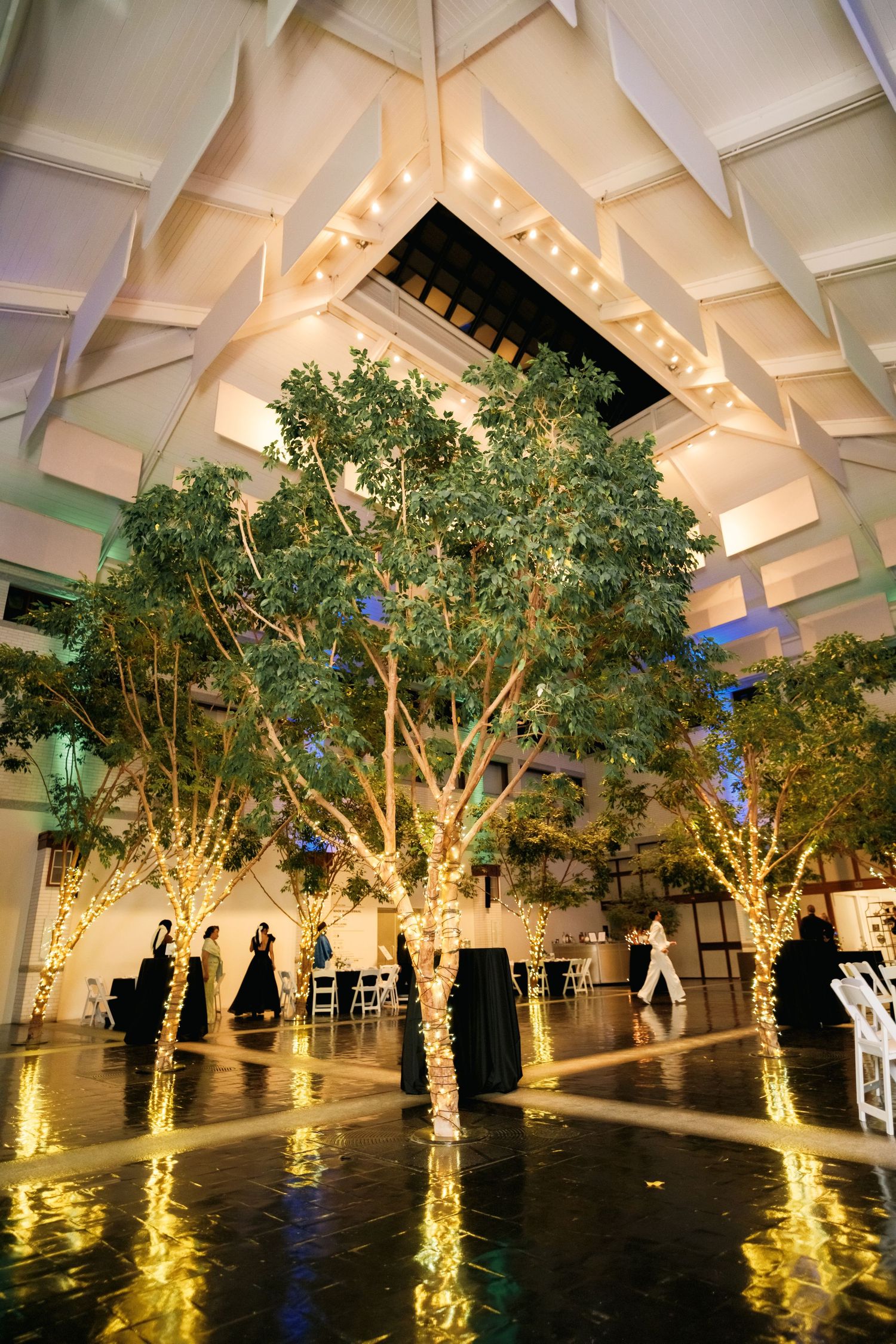 Illuminated trees with string lights create a magical indoor event space with geometric ceiling and reflective floor.