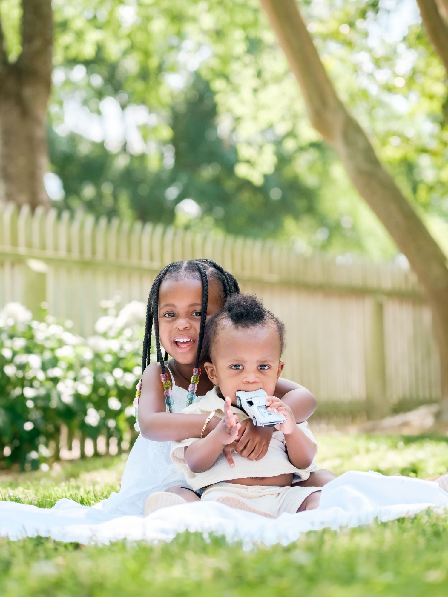 Two young siblings share playful moments together while lying on a blanket in a sunny backyard garden setting.