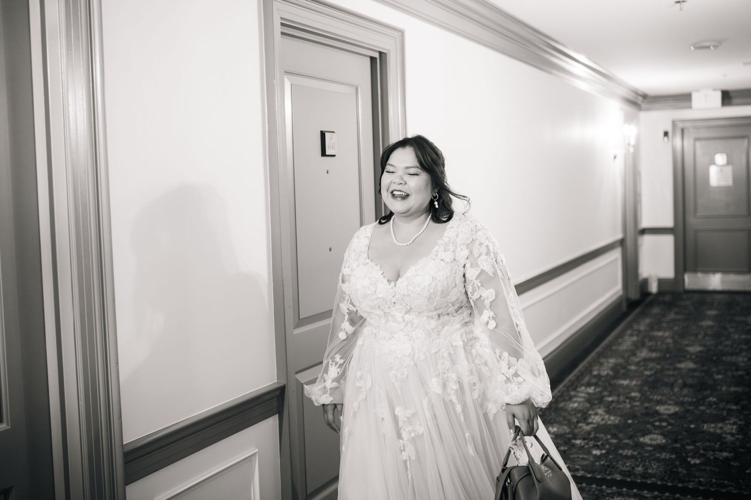 A joyful person wearing an elegant white lace wedding dress smiles while walking through a hotel hallway.