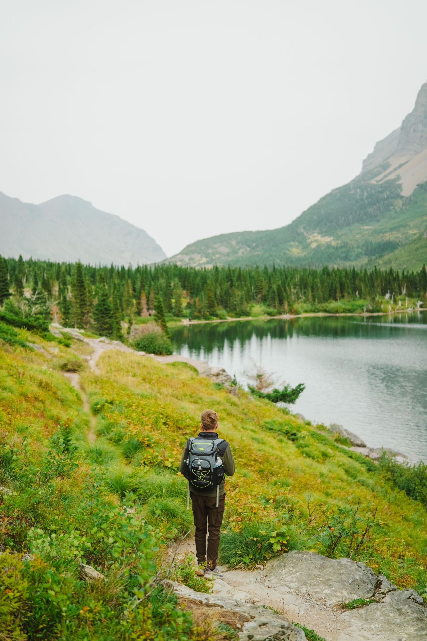 Hiker walking on scenic mountain trail along pristine lake with towering peaks and evergreen forest in background.