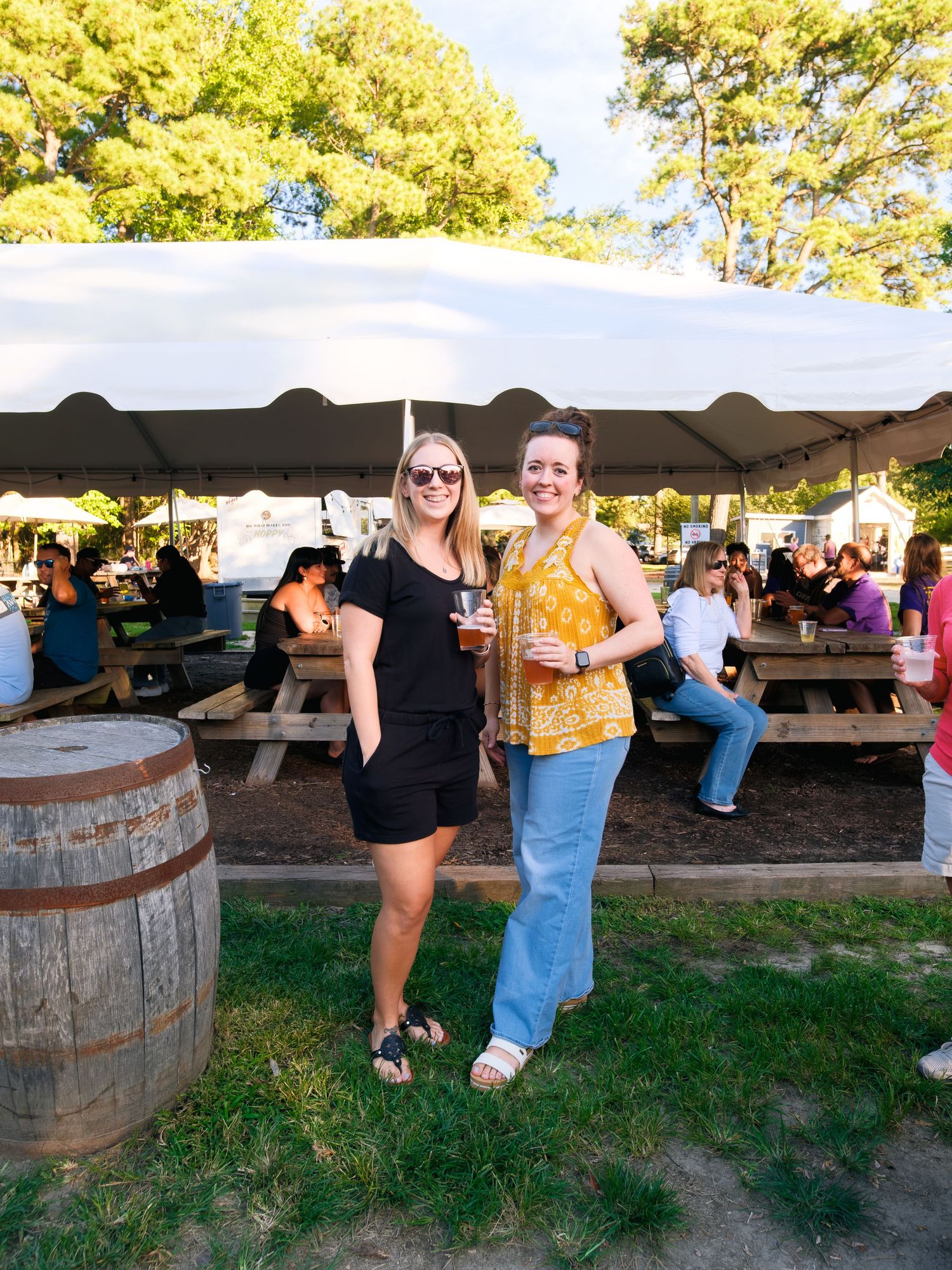 People enjoying drinks and socializing at an outdoor beer garden with wooden barrel tables at sunset.