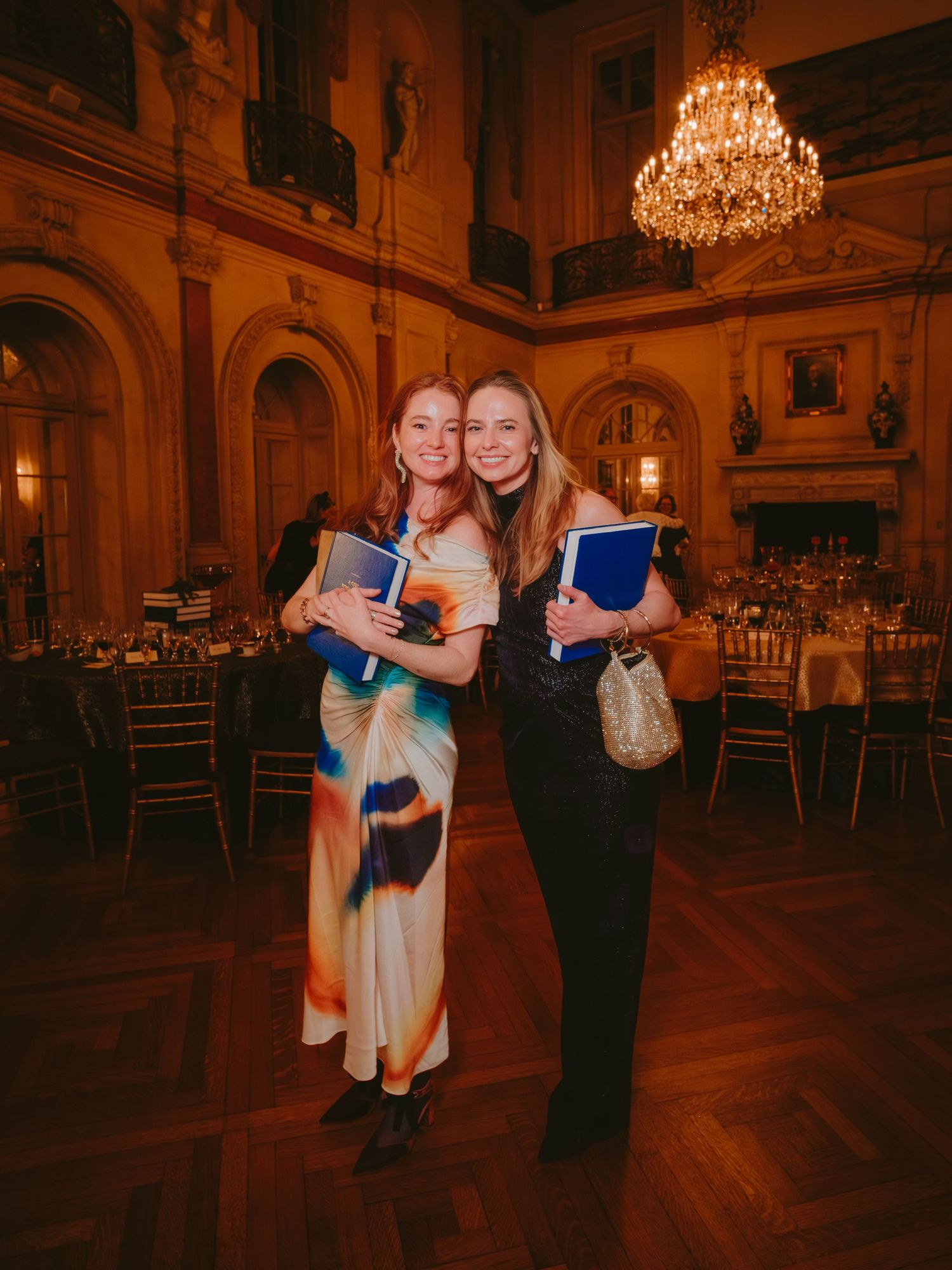Two people dance together in an elegant ballroom with ornate chandeliers and hardwood floors.